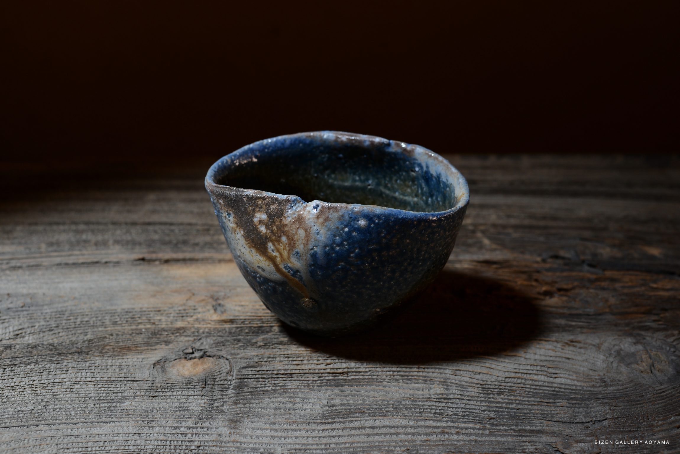 A handmade ceramic bowl with a blue and brown glaze, featuring an irregular edge, placed on a wooden surface.