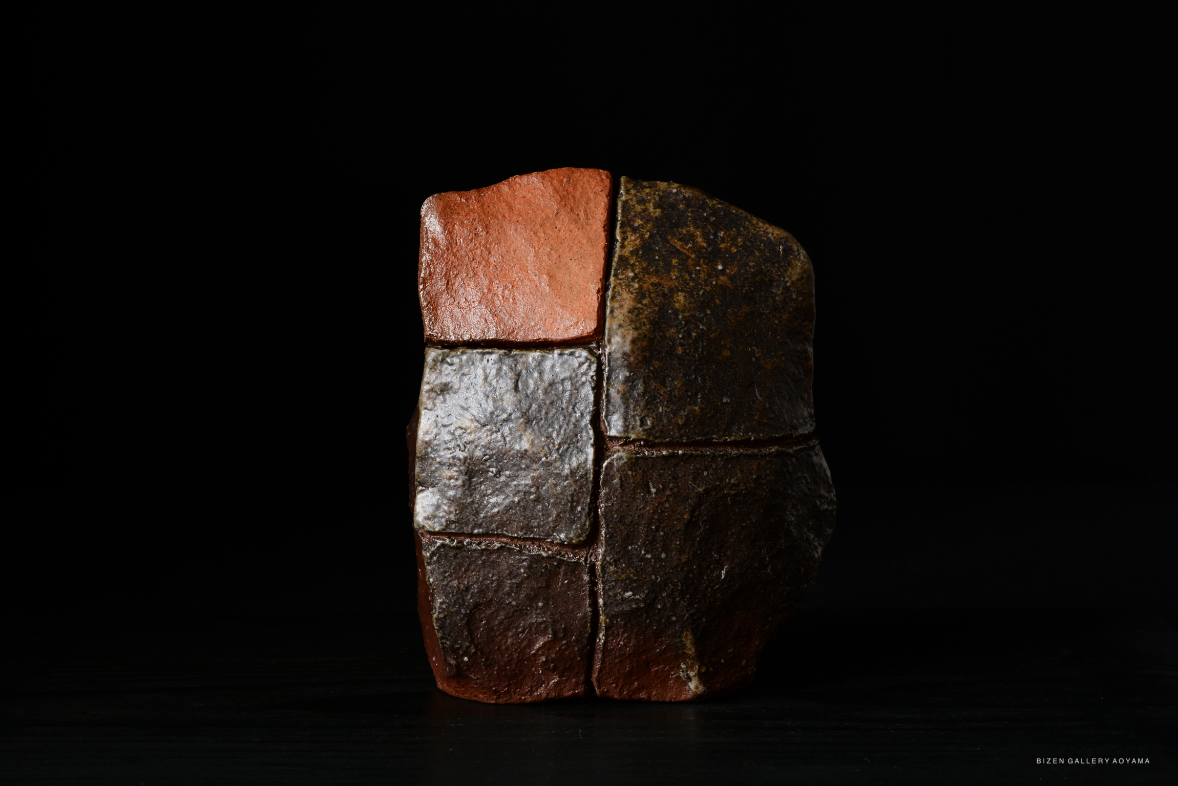 A sculptural piece resembling a block of stone divided into three distinct sections, with varying textures and colors, displayed against a black background.