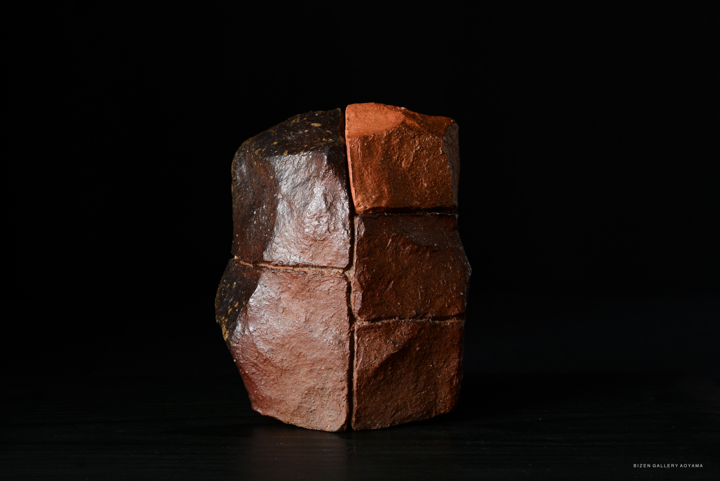 A sculptural piece made of clay with a rich red-brown color, featuring a geometric shape with multiple facets and a textured surface, set against a dark background.