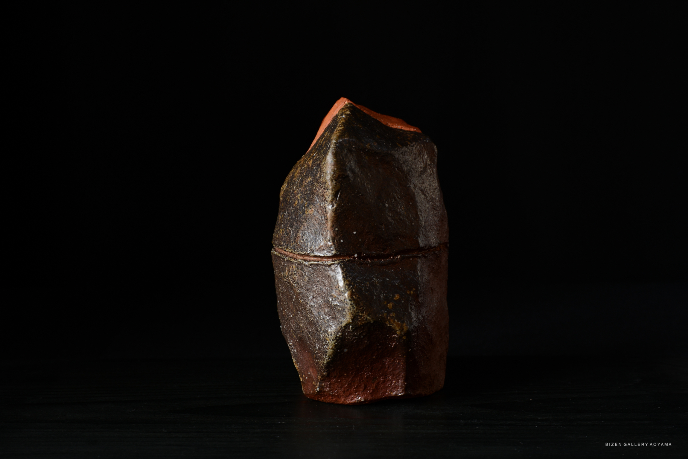 A close-up view of a unique, irregularly shaped ceramic piece with a dark, textured surface and a contrasting reddish top, set against a black background.