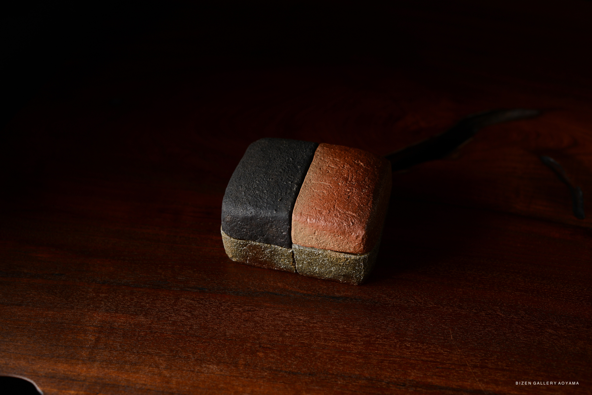 A unique piece of pottery with a two-tone design, featuring a dark surface on one side and a reddish-brown surface on the other, resting on a wooden surface.