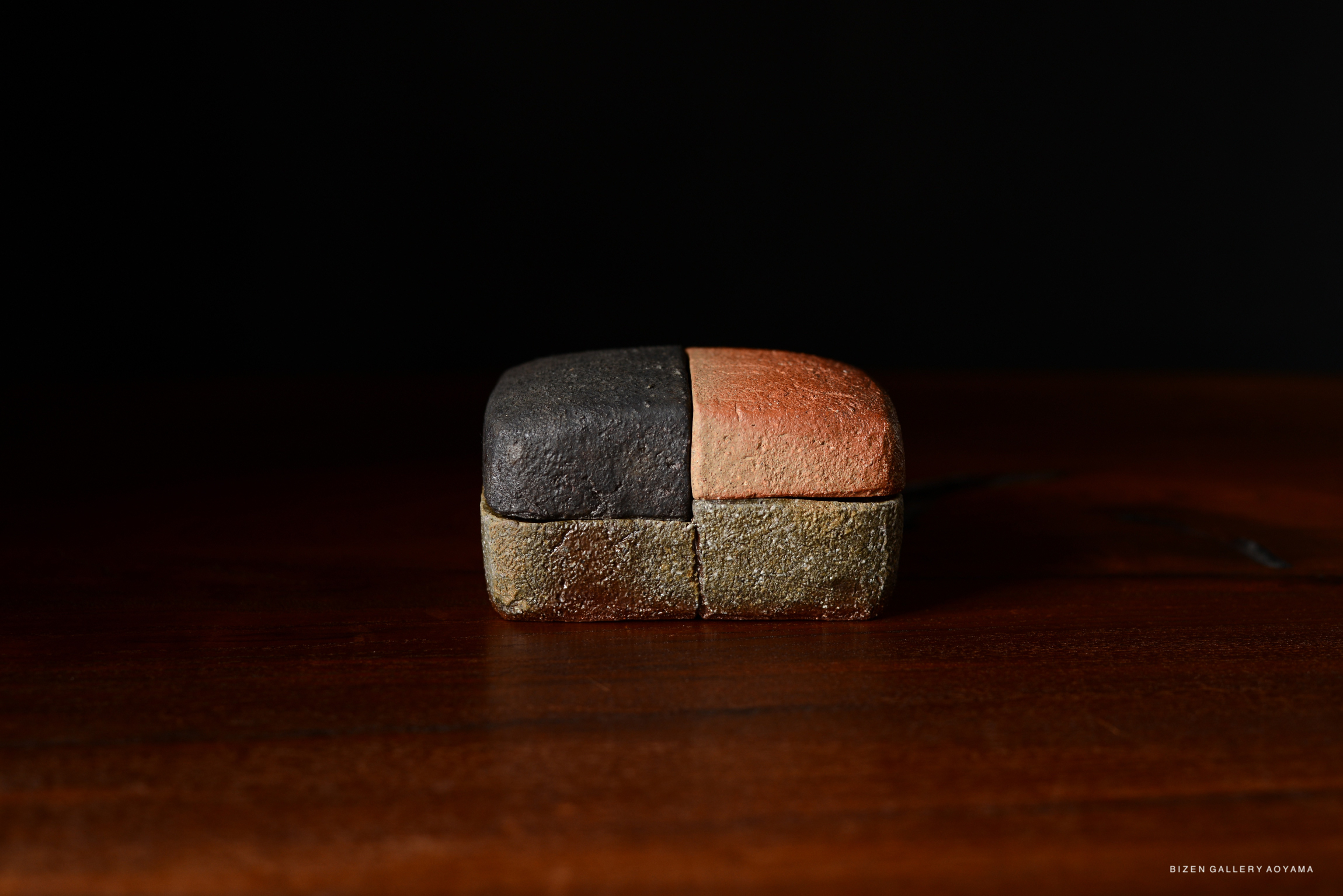 A small, rectangular Bizen pottery piece with a textured surface, featuring three distinct colors: black, reddish, and natural earthy tones. It is displayed on a wooden surface against a dark background.
