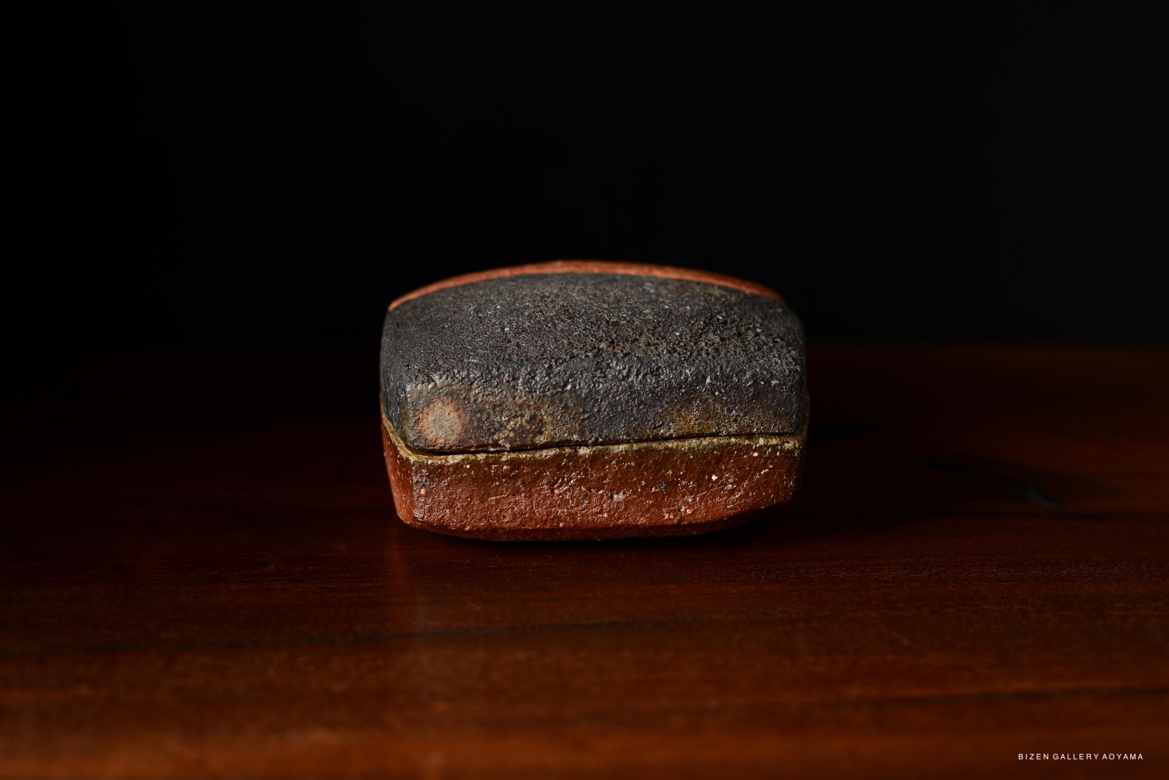 A ceramic box with a textured dark surface and a reddish-brown base, resting on a wooden table against a black background.