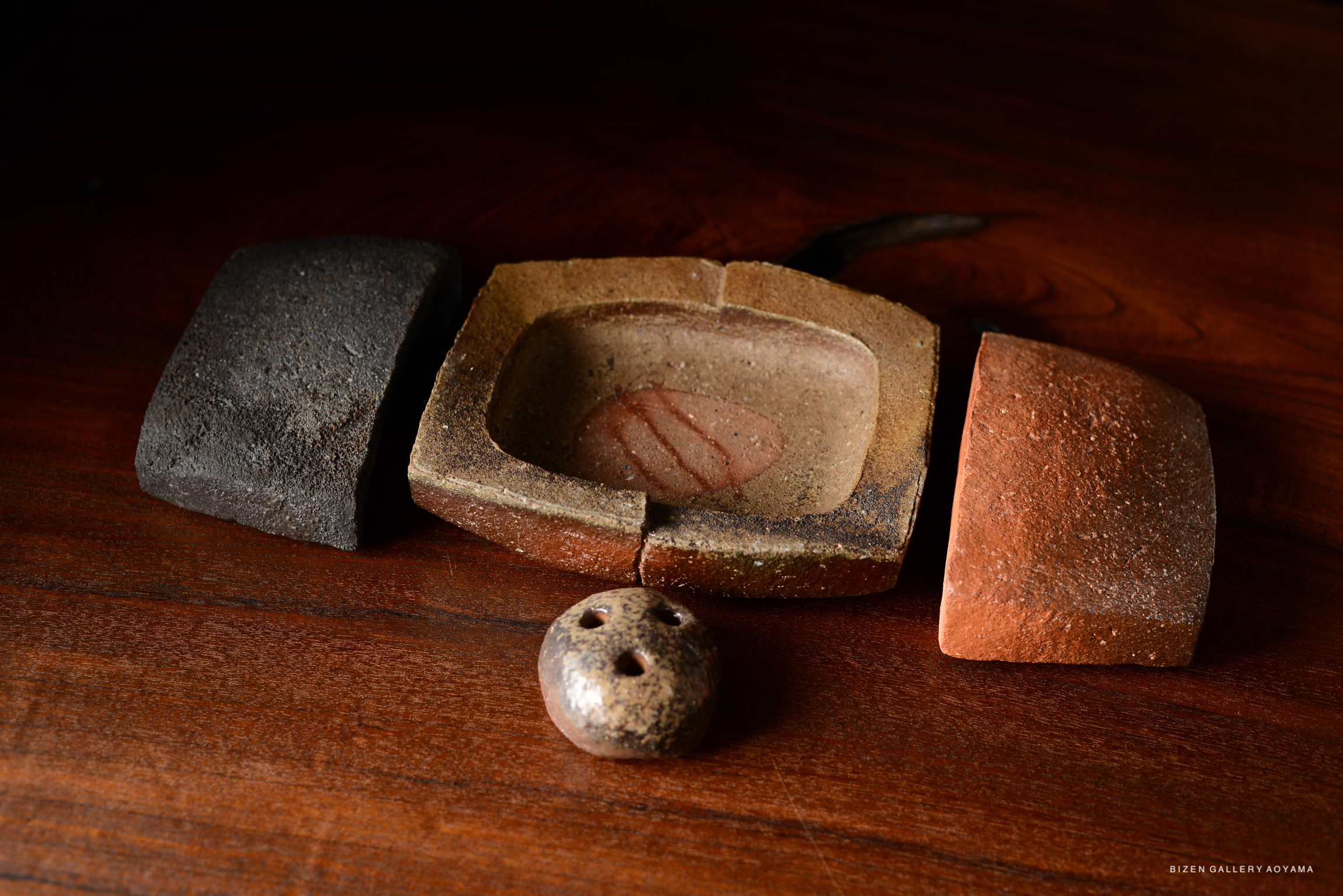 A wooden surface displaying a rustic pottery item with three pieces in various colors—dark gray, reddish-brown, and lighter brown—alongside a small round decoration showing holes.
