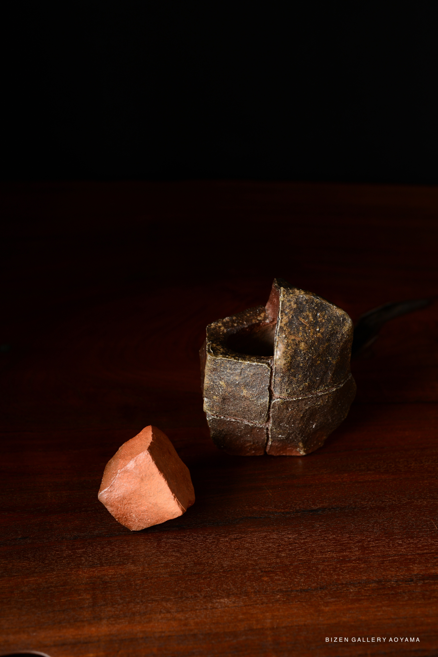 A textured dark pottery piece with a small red clay stone beside it, set against a black background.