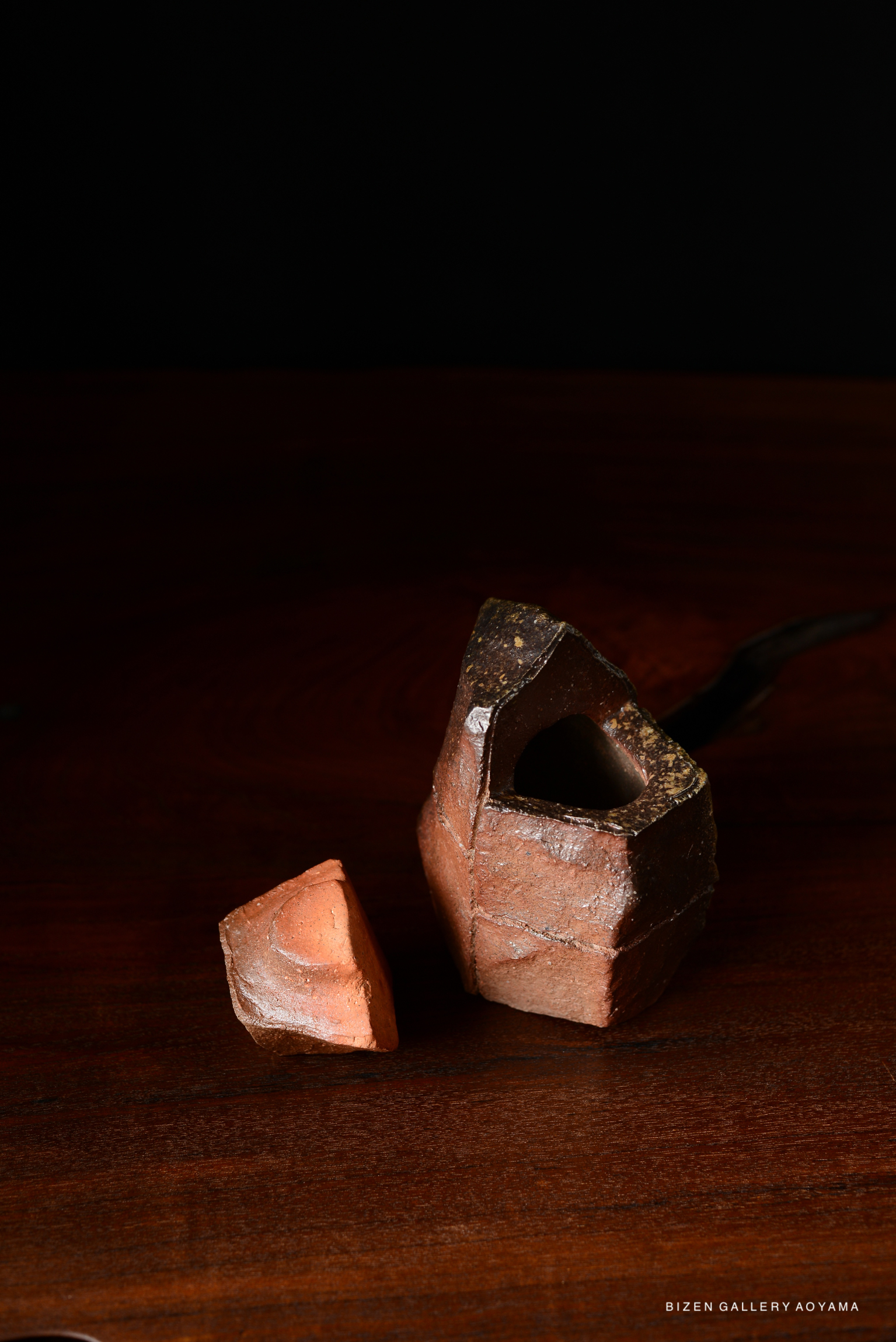 A close-up of two pieces of pottery, one with an open shape and intricate surface details, resting on a wooden surface against a dark background.