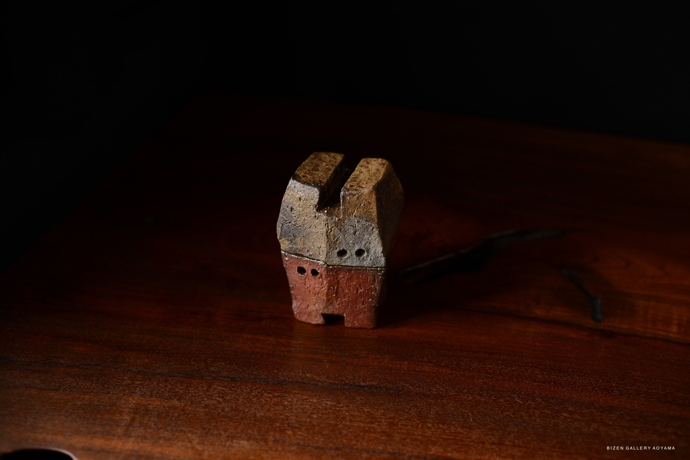A small ceramic sculpture resembling a house, featuring a gray and reddish-brown color scheme, displayed on a wooden surface.