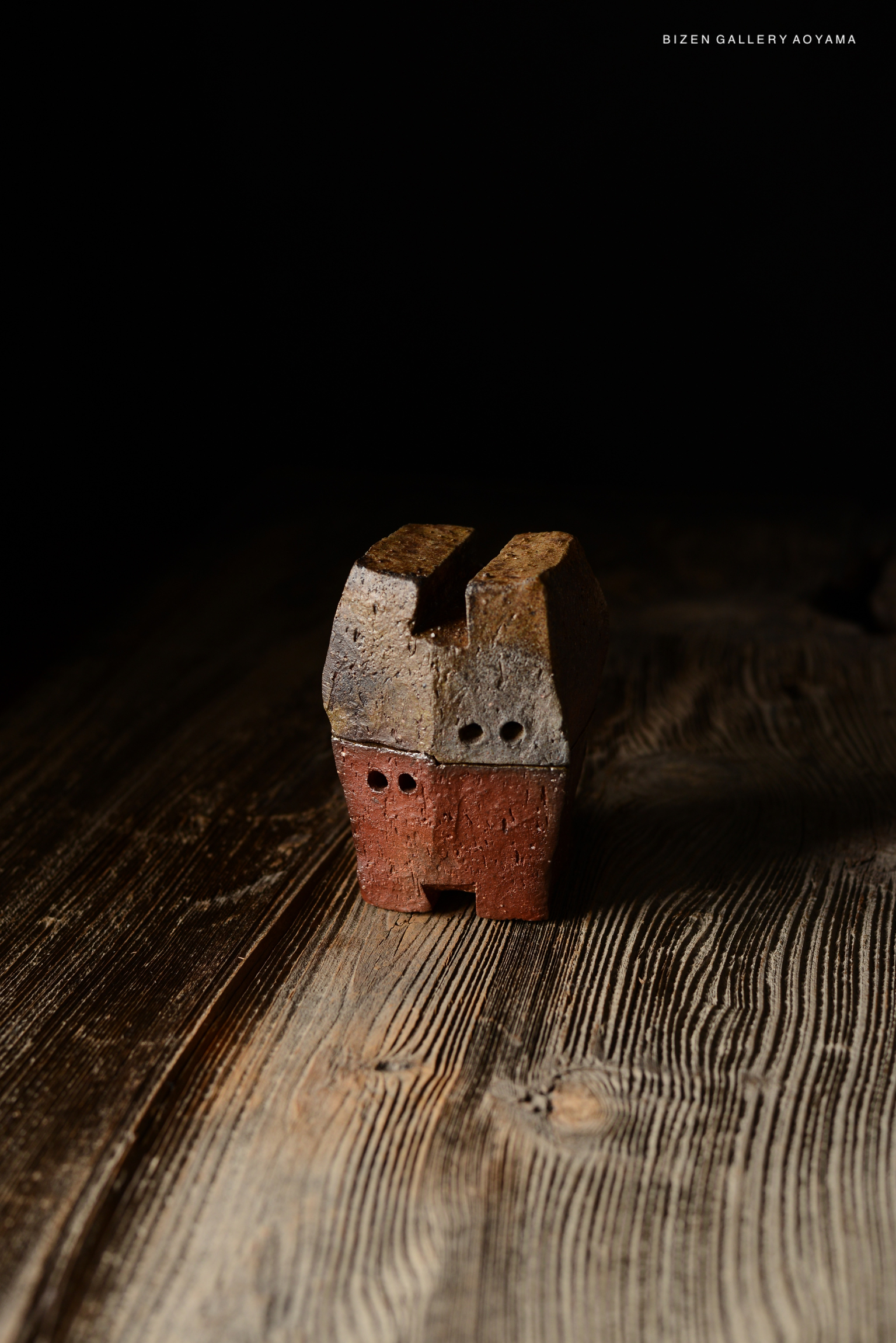 A small, artistic pottery sculpture resembling a house, made from textured clay, sitting on a wooden surface with a dark background.