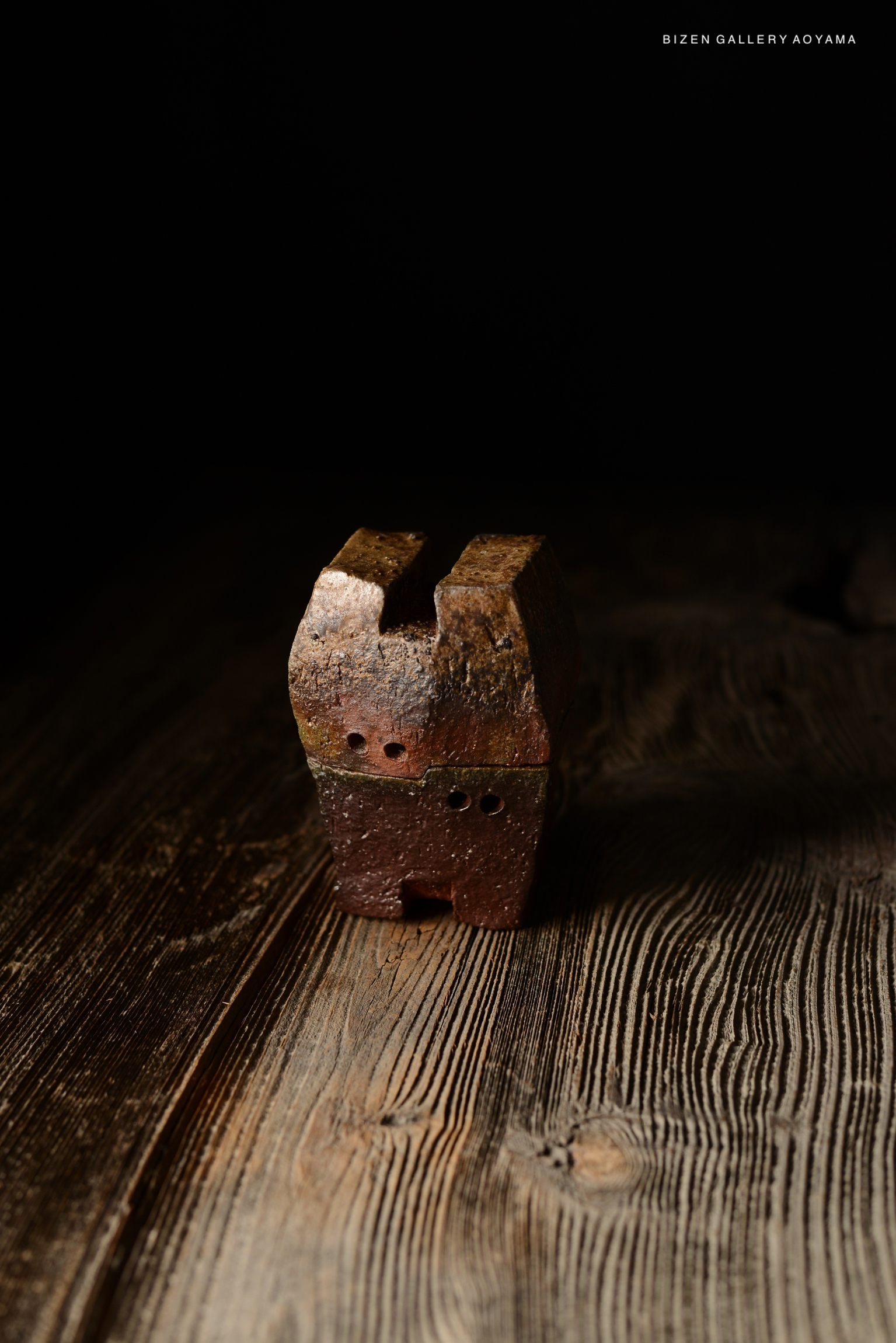 A rustic ceramic sculpture with a unique shape, featuring holes and a textured surface, on a wooden table with a dark background.