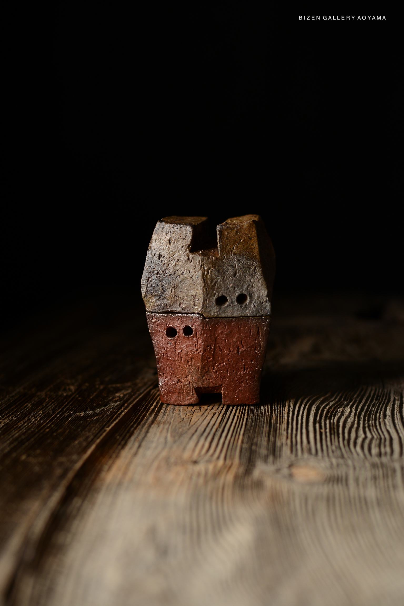 A small, unique ceramic sculpture with a rustic texture, featuring two holes for eyes and a mouth, displayed against a dark background.