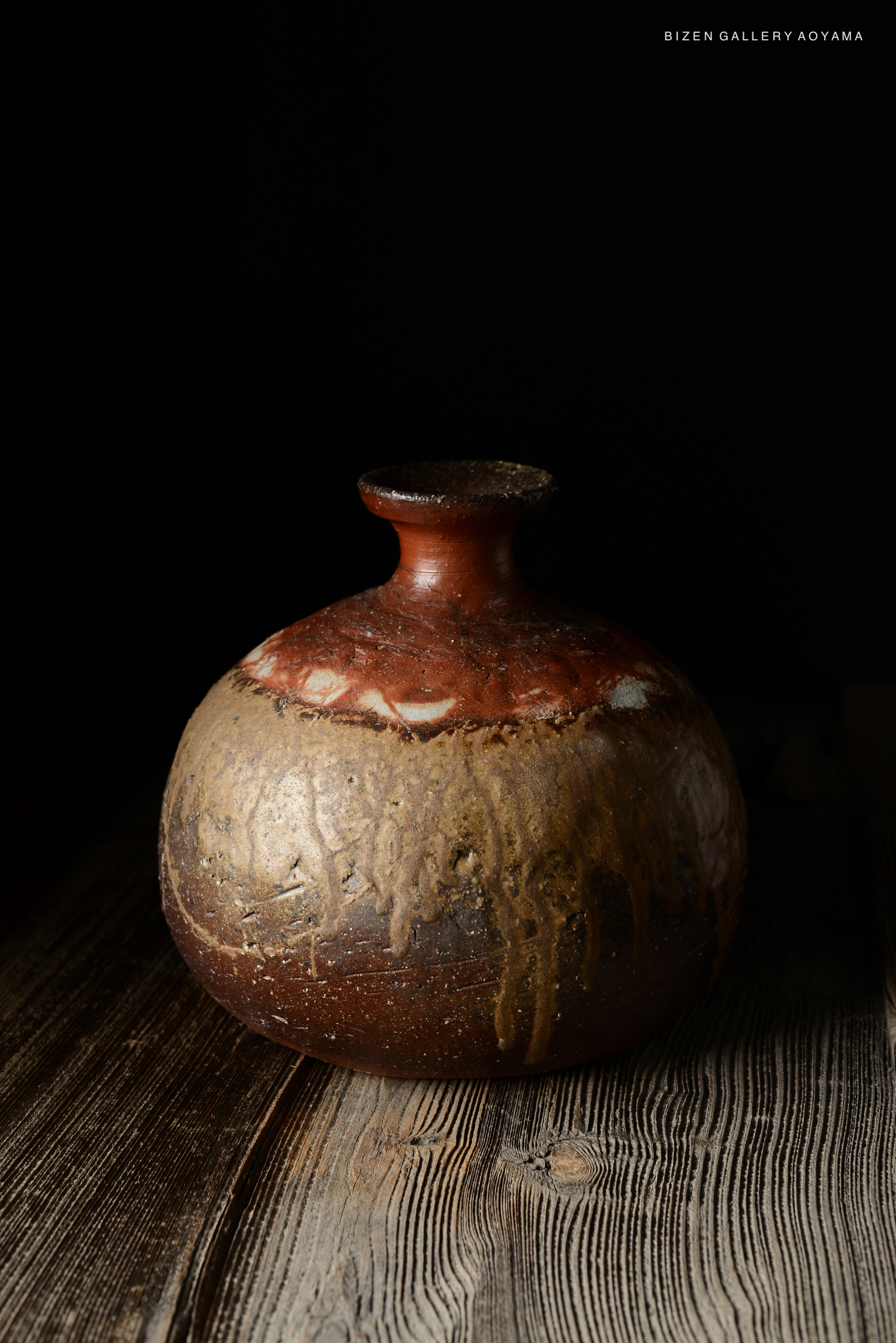 A traditional Bizen pottery Tokkuri (sake bottle) with a distinctive round body and narrow neck, featuring a combination of earthy colors and natural glaze drippings, displayed on a wooden surface.