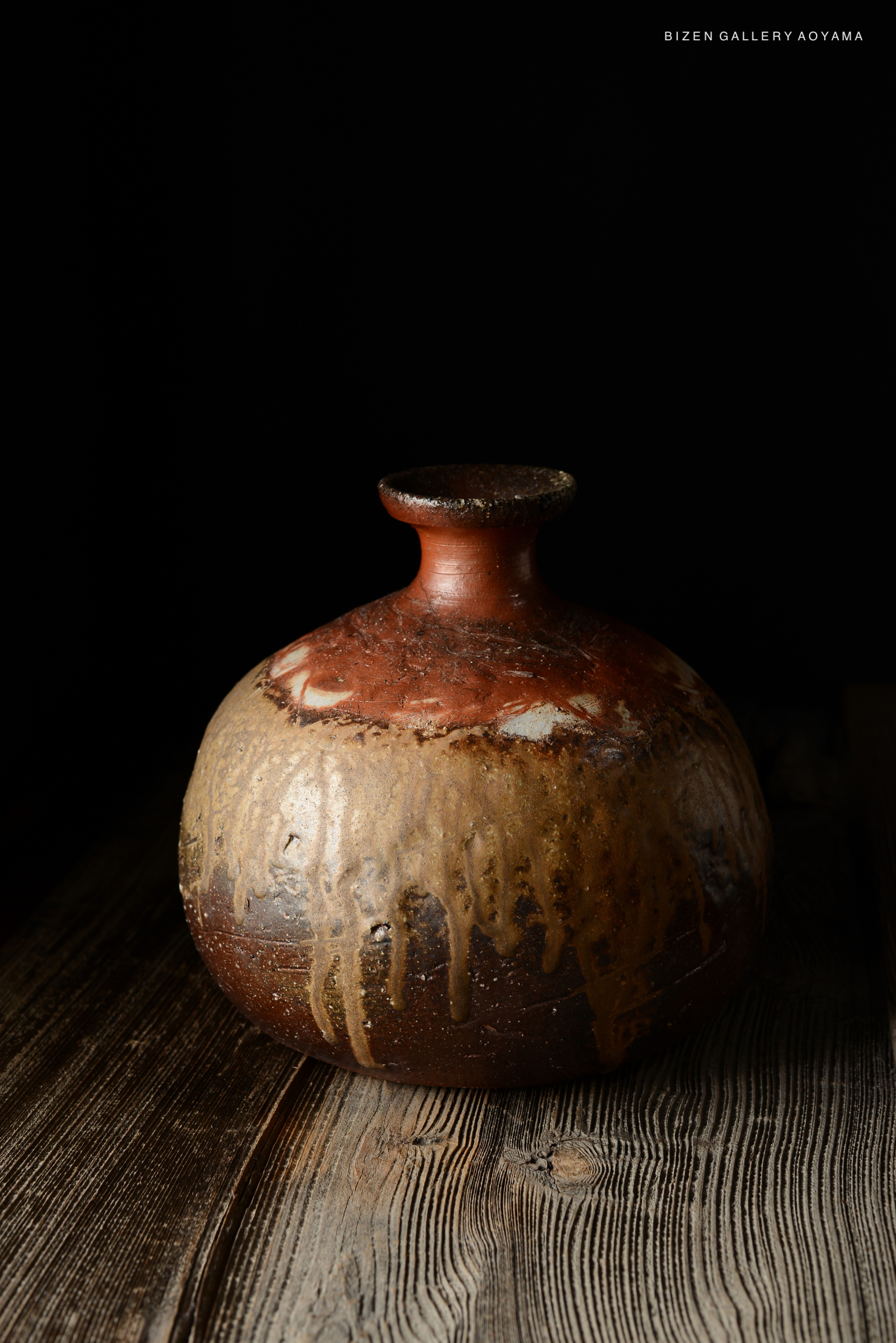 A Bizen O Kabura Tokkuri styled ceramic sake bottle with a rounded body and a narrow neck, exhibiting a mixture of earthy colors and drips of glaze, set against a dark background and wooden surface.