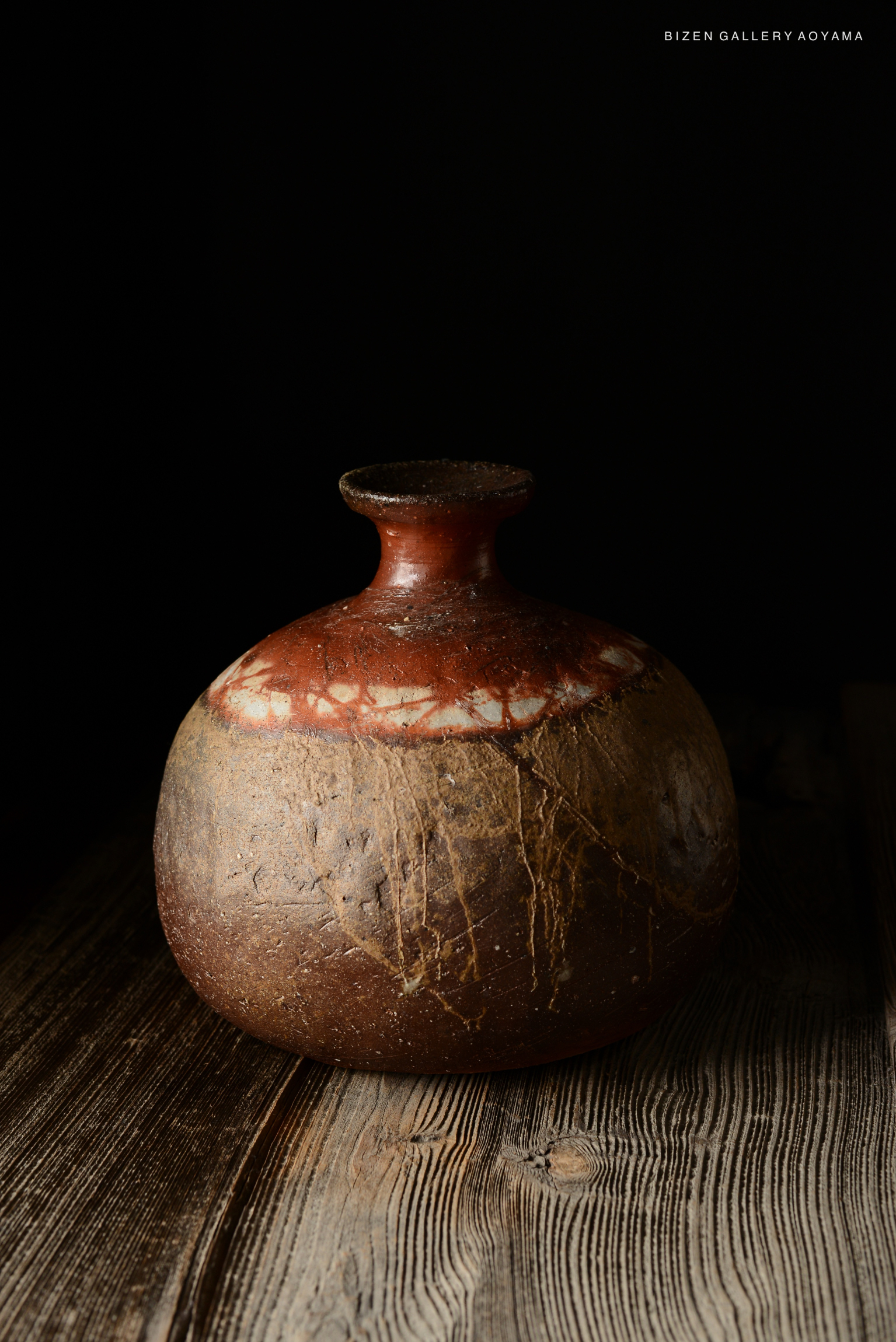 A textured Bizen pottery tokkuri (sake flask) with a rounded body and narrow neck, featuring earthy tones and a distinctive glaze pattern, placed on a wooden surface.