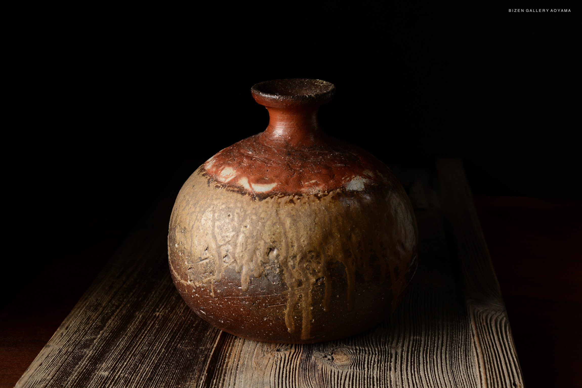 A close-up view of a round Bizen pottery Tokkuri with a rustic glaze, resting on a wooden surface against a dark background.