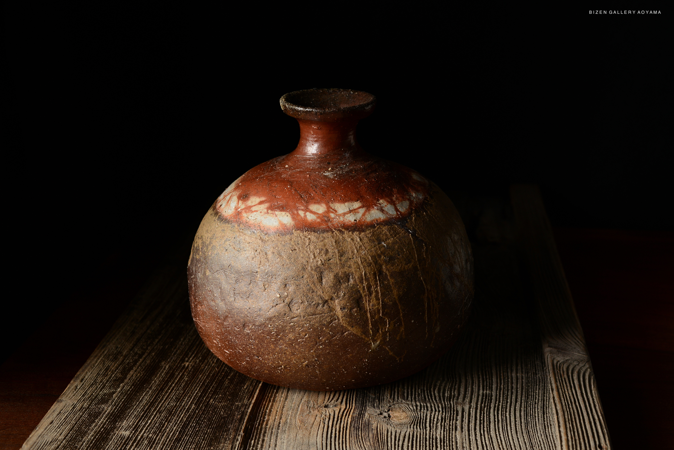 A Bizen pottery Tokkuri (sake vessel) with a rounded body and a narrow neck, showcasing natural earthy tones and textures, placed on a wooden surface against a dark background.