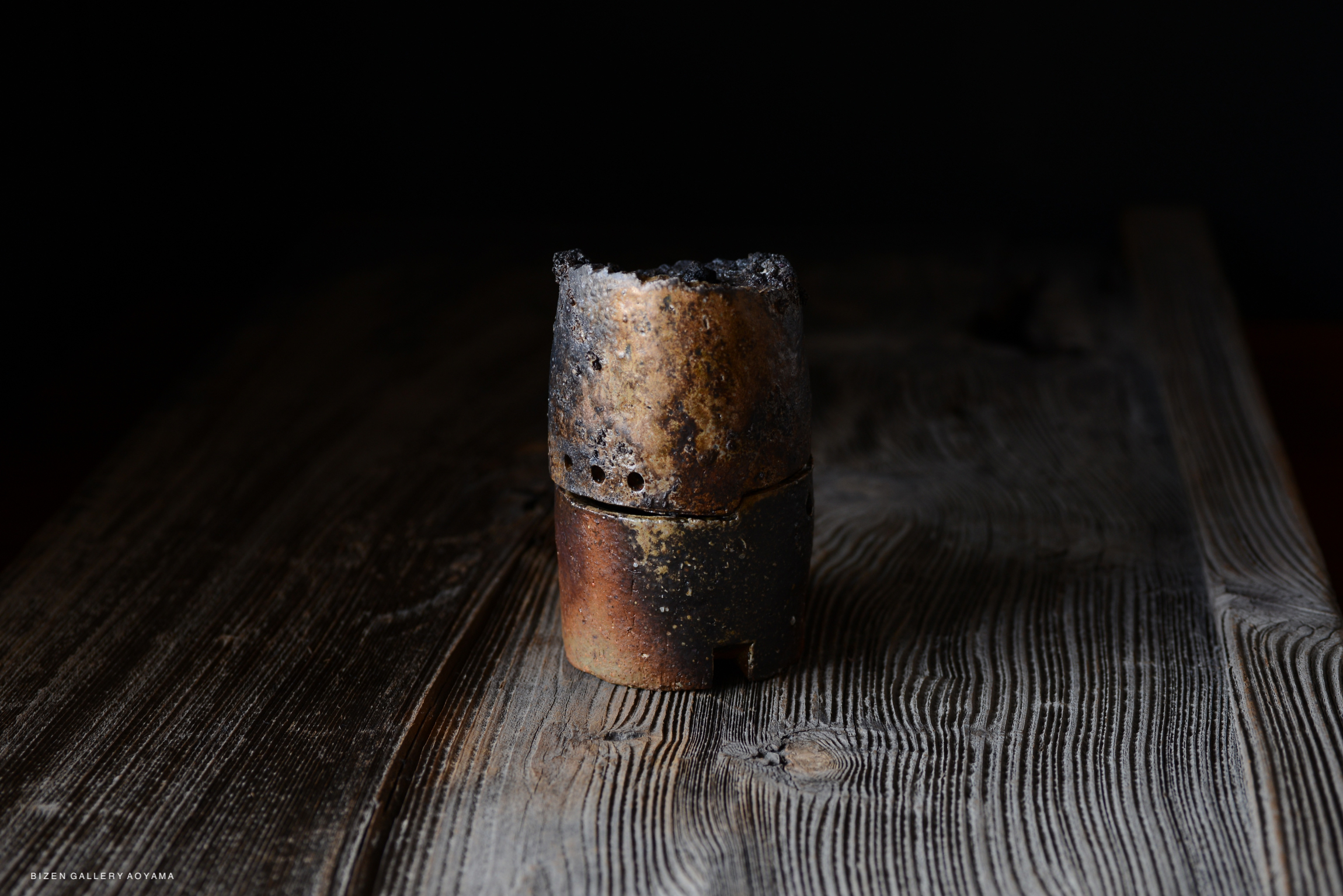 A rustic, cylindrical ceramic piece with a charred top, sitting on a textured wooden surface, against a dark background.