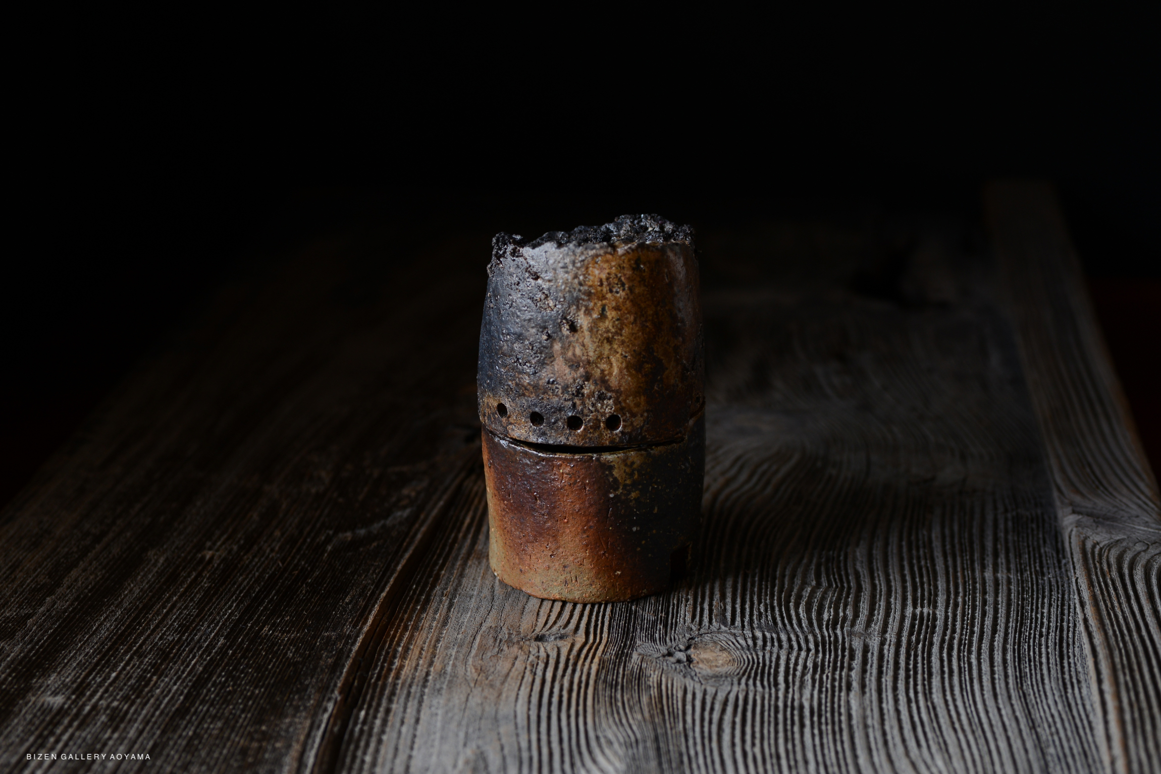 A small, cylindrical pottery piece with a dark, textured surface, partially burnt and sitting on a wooden surface.