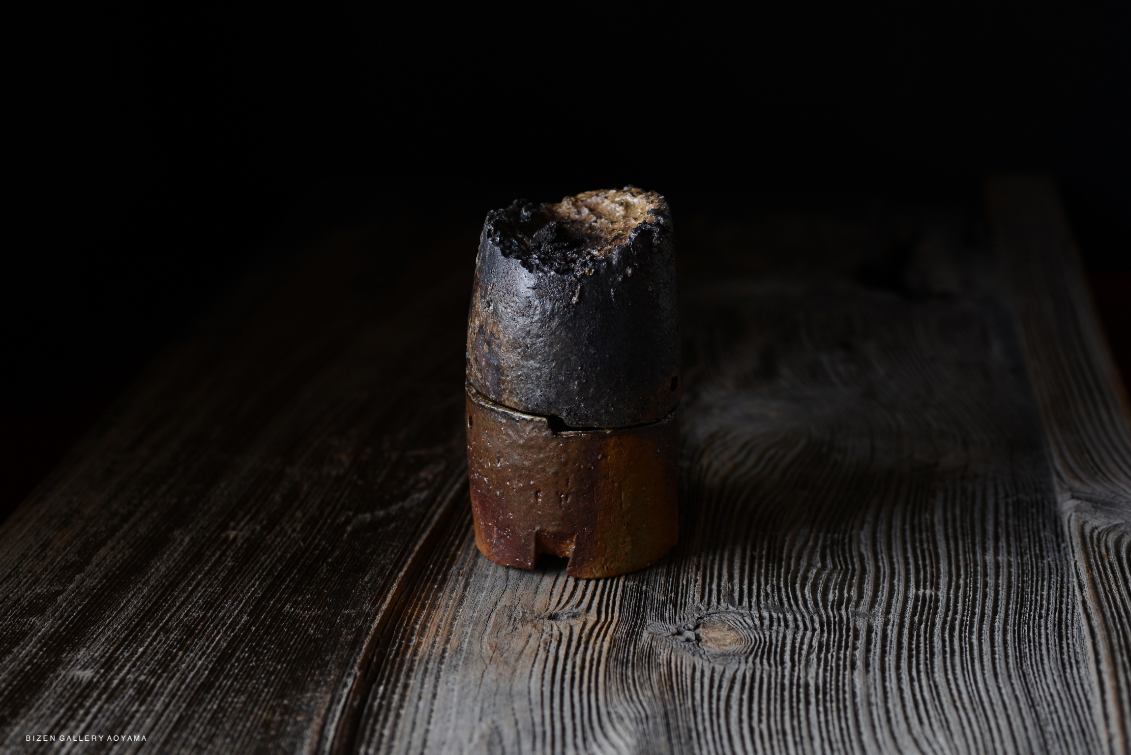 A piece of pottery with a blackened top and reddish-brown base, placed on a textured wooden surface.