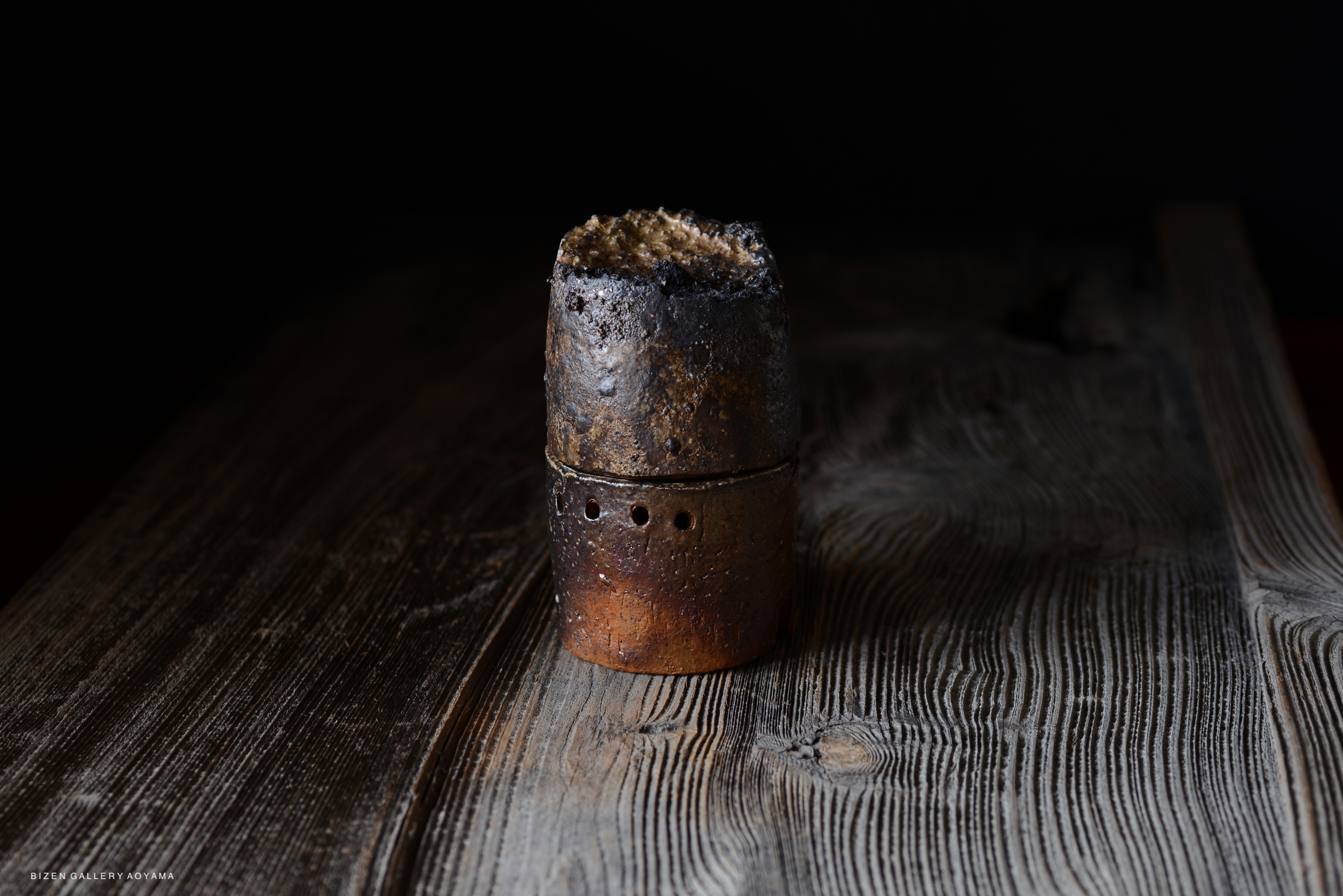A handcrafted ceramic piece with a textured blackened top, resting on a rustic wooden surface.