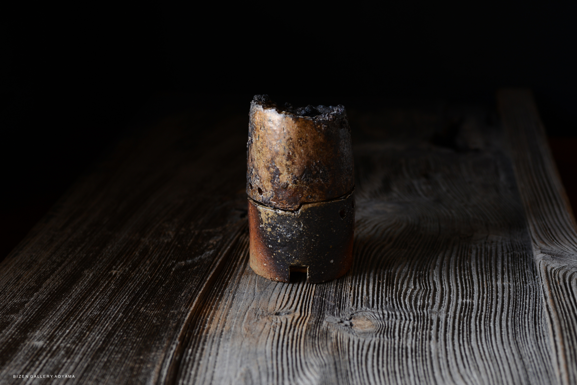 A rustic, handmade Bizen pottery vessel displayed on a wooden surface, featuring a textured, darkened exterior.
