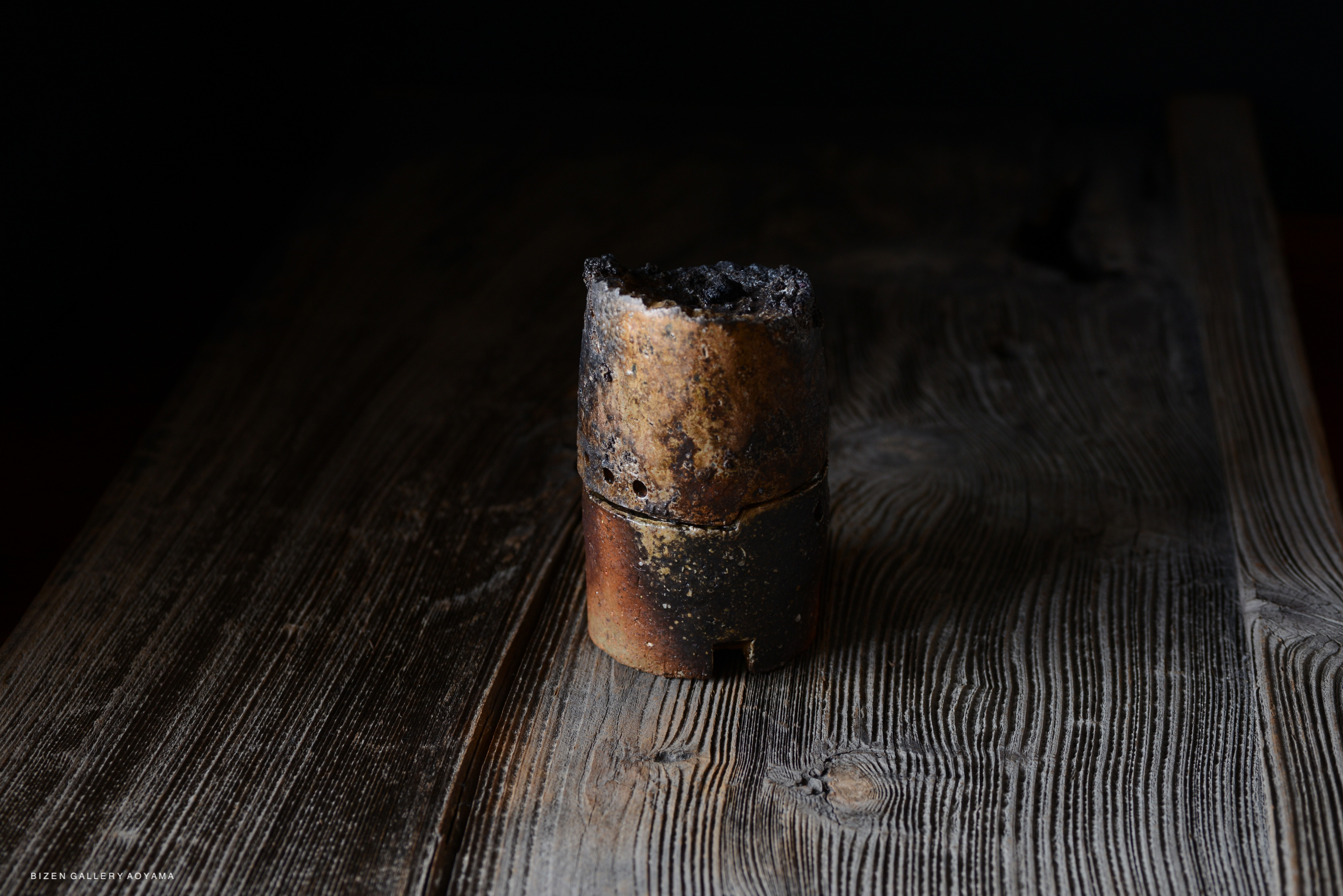 A ceramic vessel with a dark, burnished surface, showcasing uneven textures and colors, placed on a wooden surface.