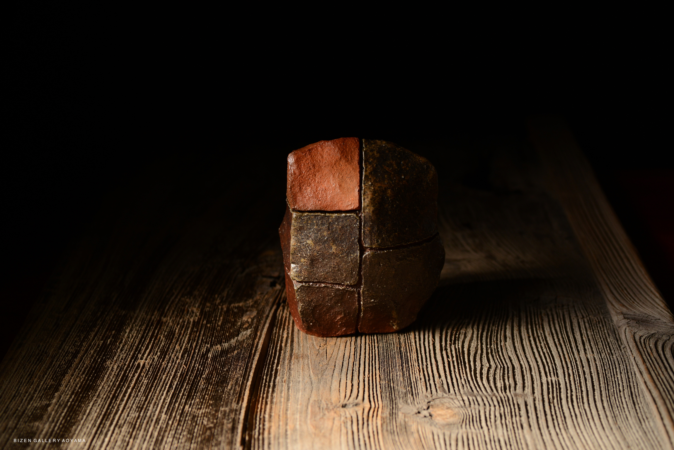 A textured, irregularly shaped ceramic piece with brown and black sections, resting on a wooden surface against a dark background.