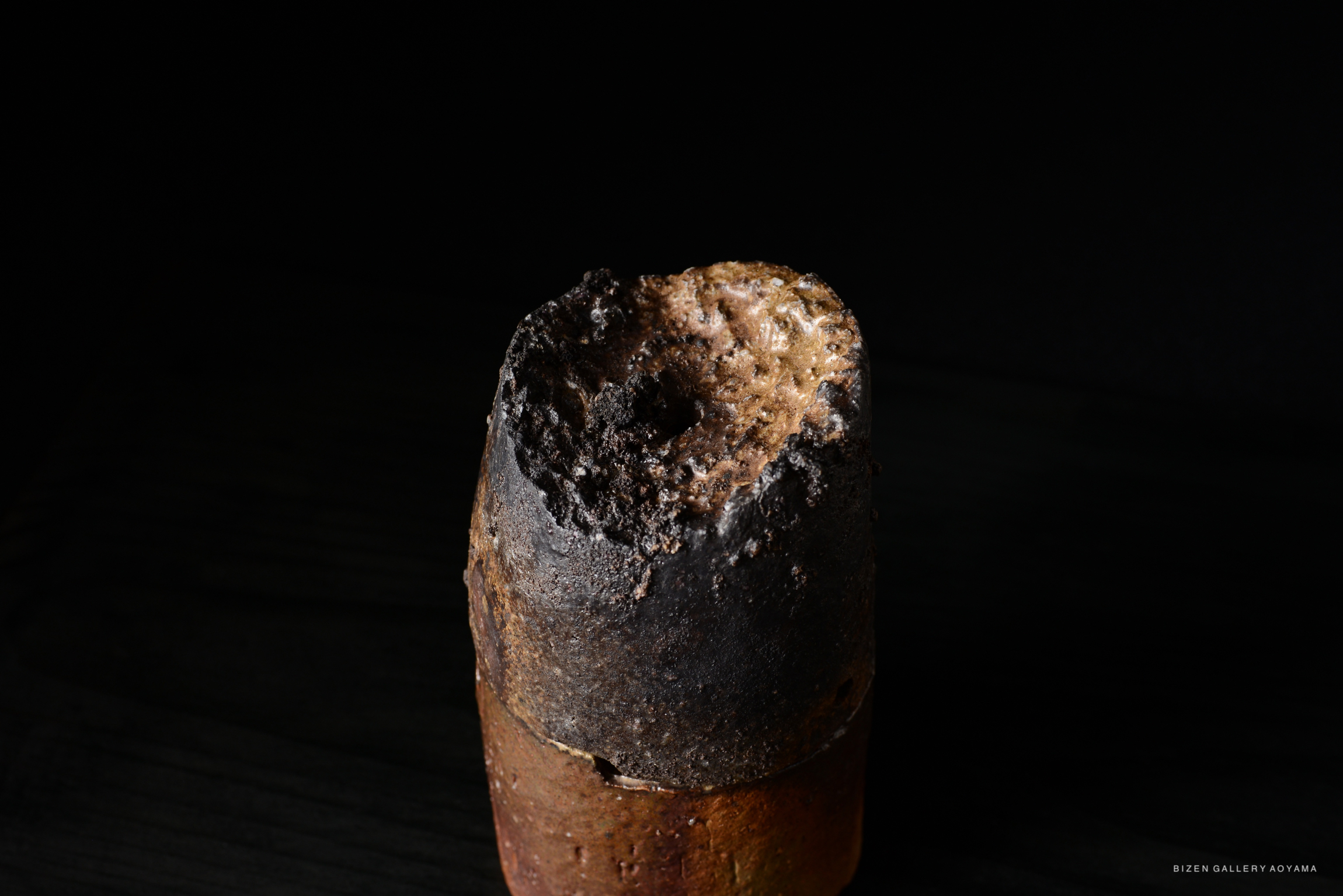 Close-up of a Bizen pottery piece with a dark, scorched surface and a rounded, irregular top against a black background.