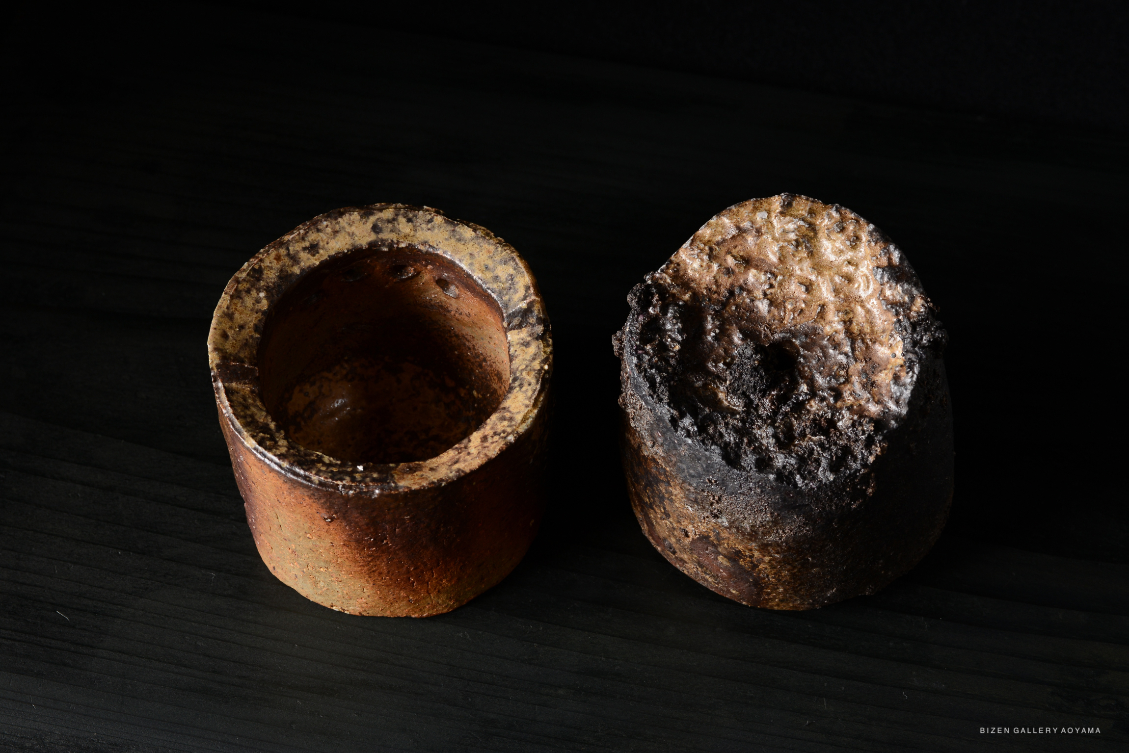 Two handcrafted ceramic pots displayed on a dark surface, one with a smooth brown interior and exterior, the other with a textured burnt top and rough blackened sides.