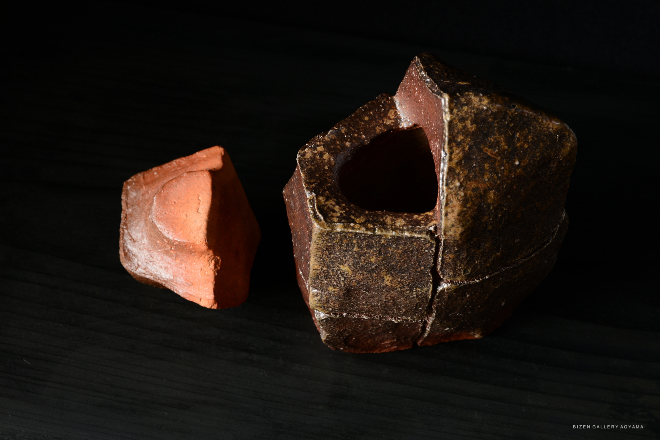 Two pieces of pottery are arranged on a dark surface, one is irregularly shaped and has a hole, while the other is smaller and more angular.
