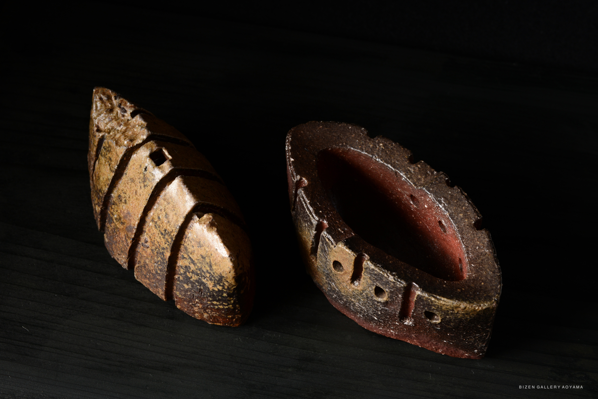 Two unique ceramic pieces resembling boats, one with a textured brown surface and the other reddish-brown, displayed on a dark background.
