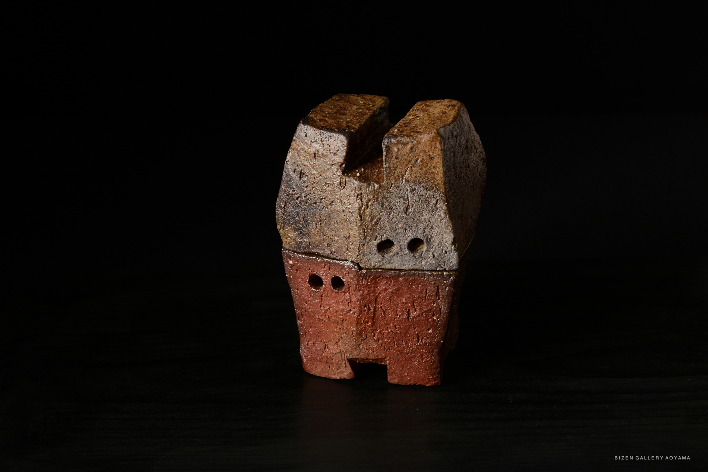 A unique, abstract ceramic sculpture featuring a combination of textured gray and reddish-brown surfaces with small openings, set against a dark background.