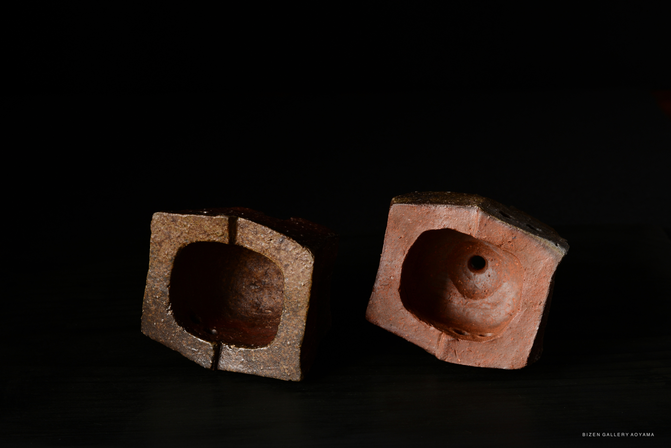 Two unique Bizen pottery pieces with a rustic finish, showcasing irregular shapes and textured surfaces against a dark background.