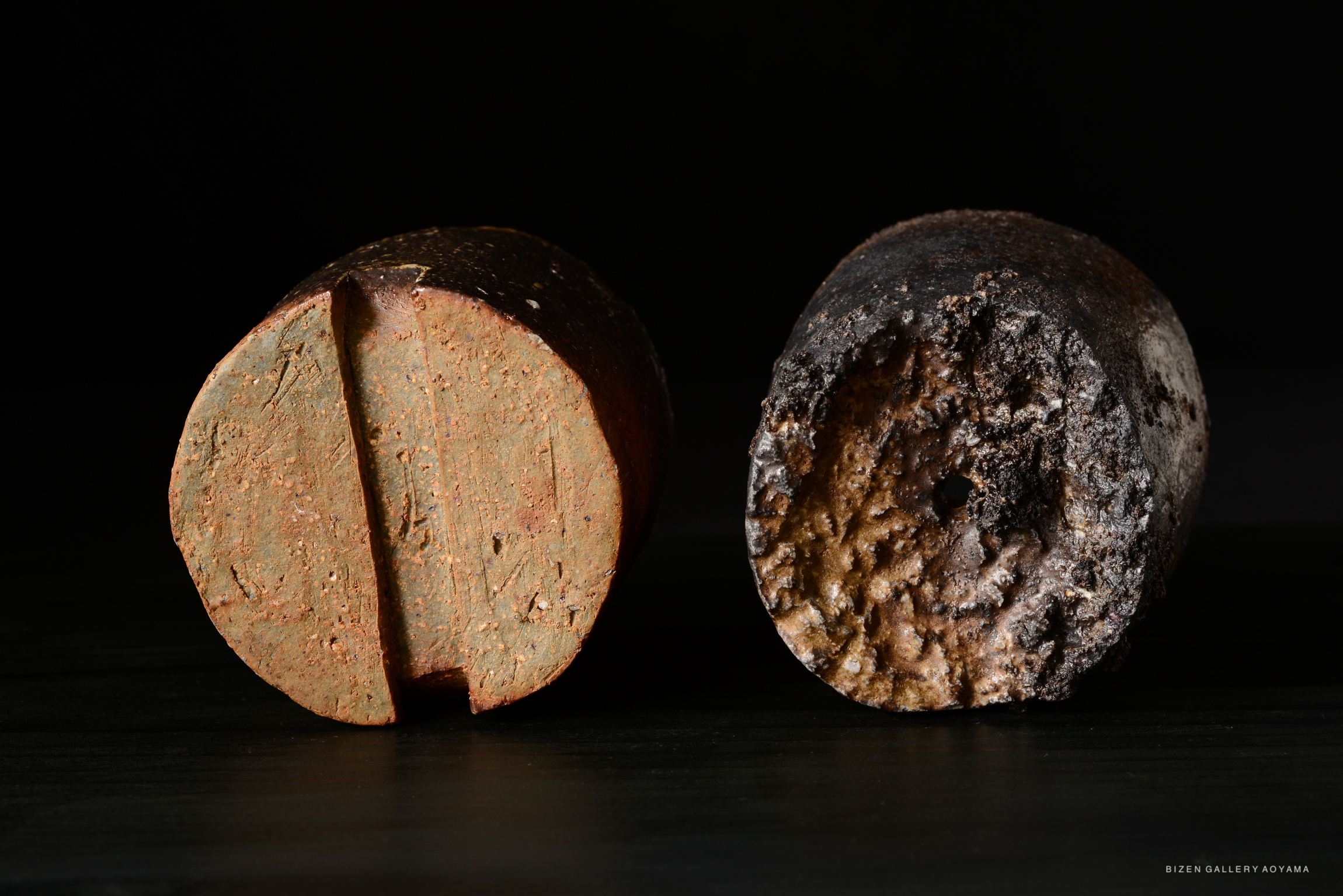 Two cylindrical pieces of pottery displayed on a dark background. The left piece has a reddish-brown color with distinct uneven textures and a shallow groove running vertically. The right piece is darker, showing a charred and rough surface with signs of aging and wear.