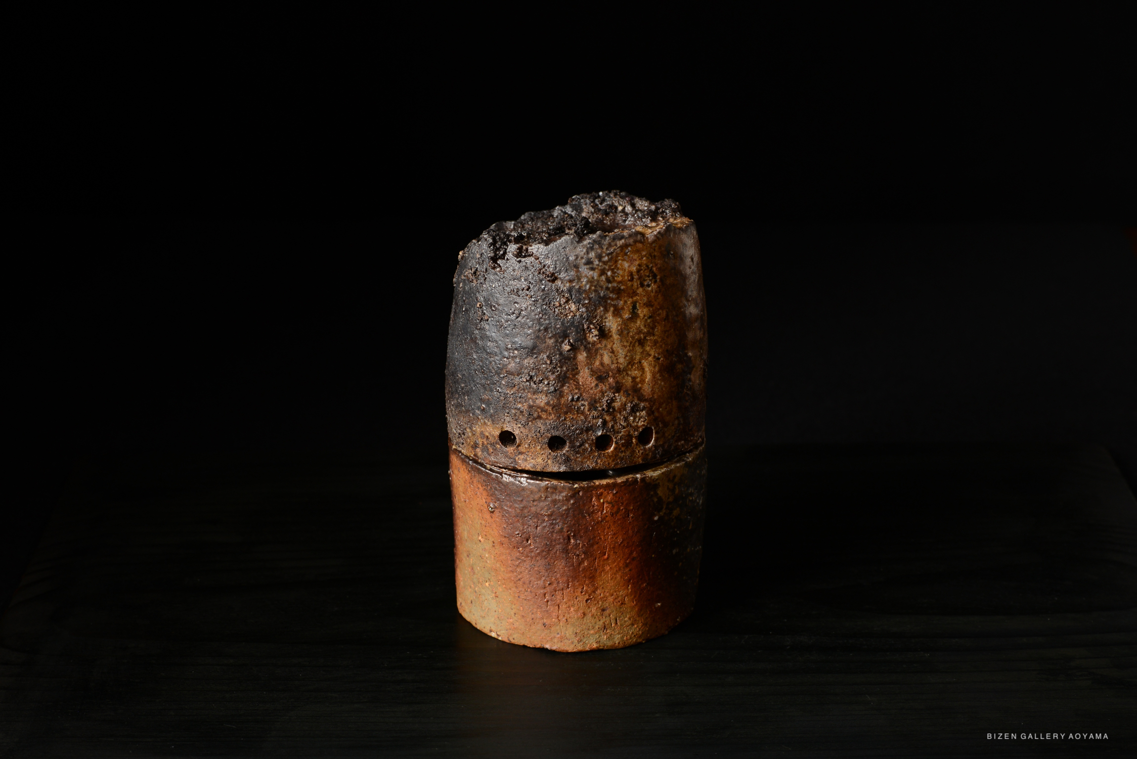 A rustic Bizen pottery piece with a charred top and a smooth, reddish-brown lower section, set against a black background.