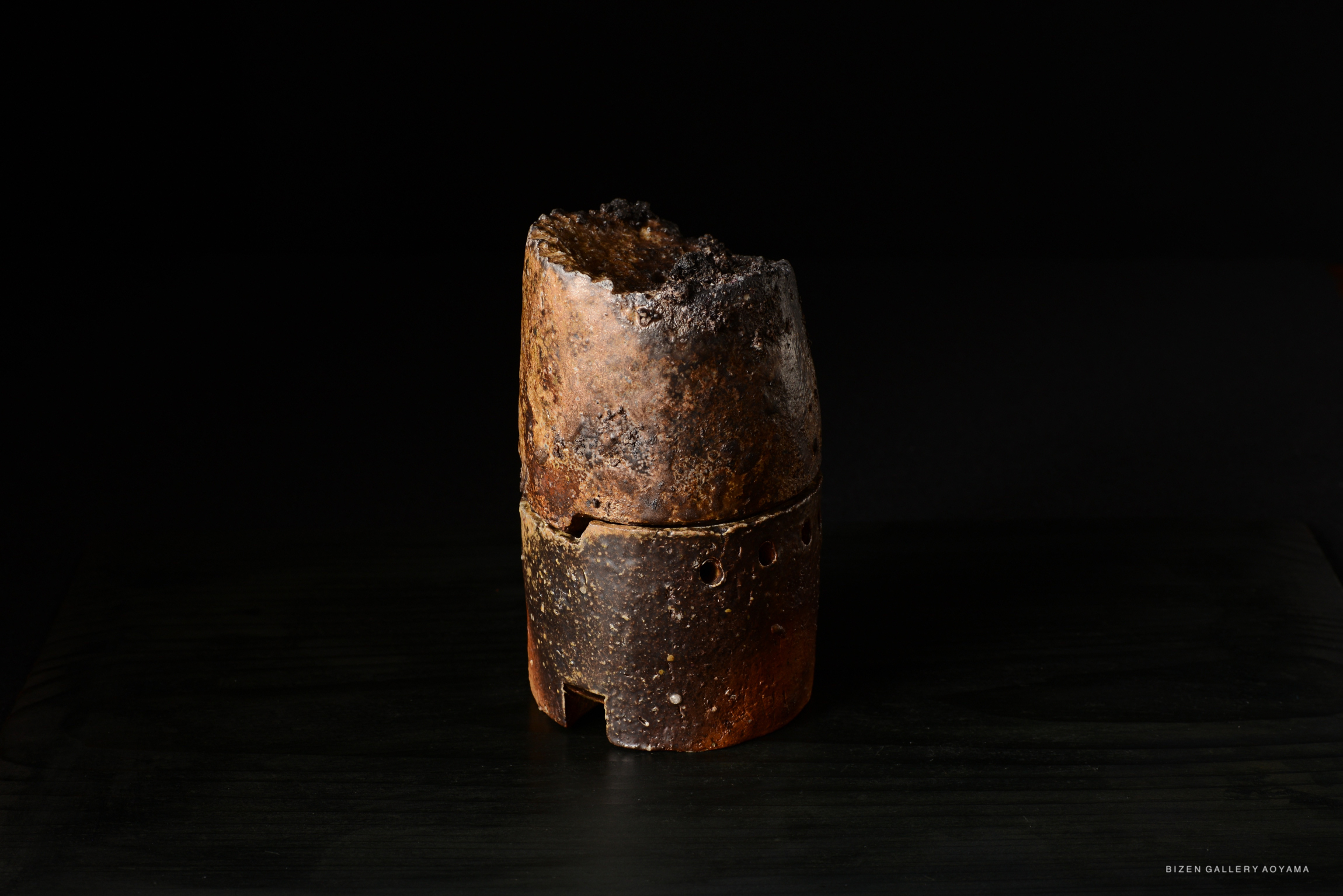 A rustic ceramic vessel with a dark, textured surface, displayed against a black background.