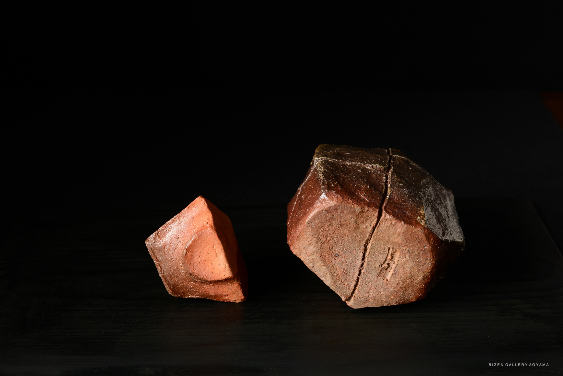 Two ceramic pieces displayed on a black surface. The larger piece on the right has a rough texture and an irregular shape, while the smaller piece on the left is triangular with a smoother finish.
