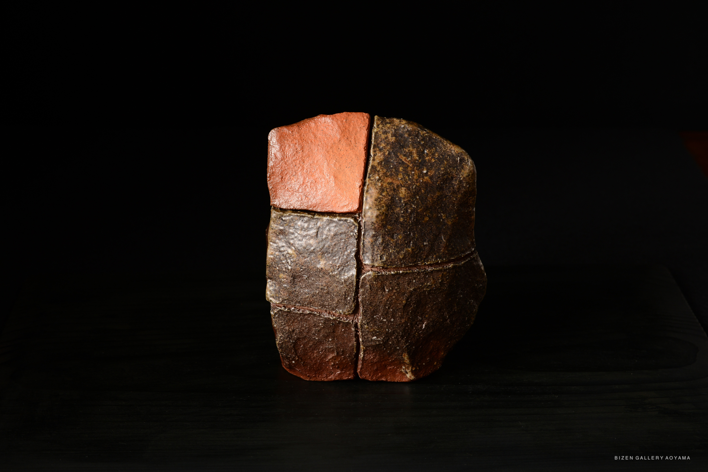 A sculptural clay object with a mix of earthy tones, featuring distinct sections and textured surfaces, displayed against a dark background.