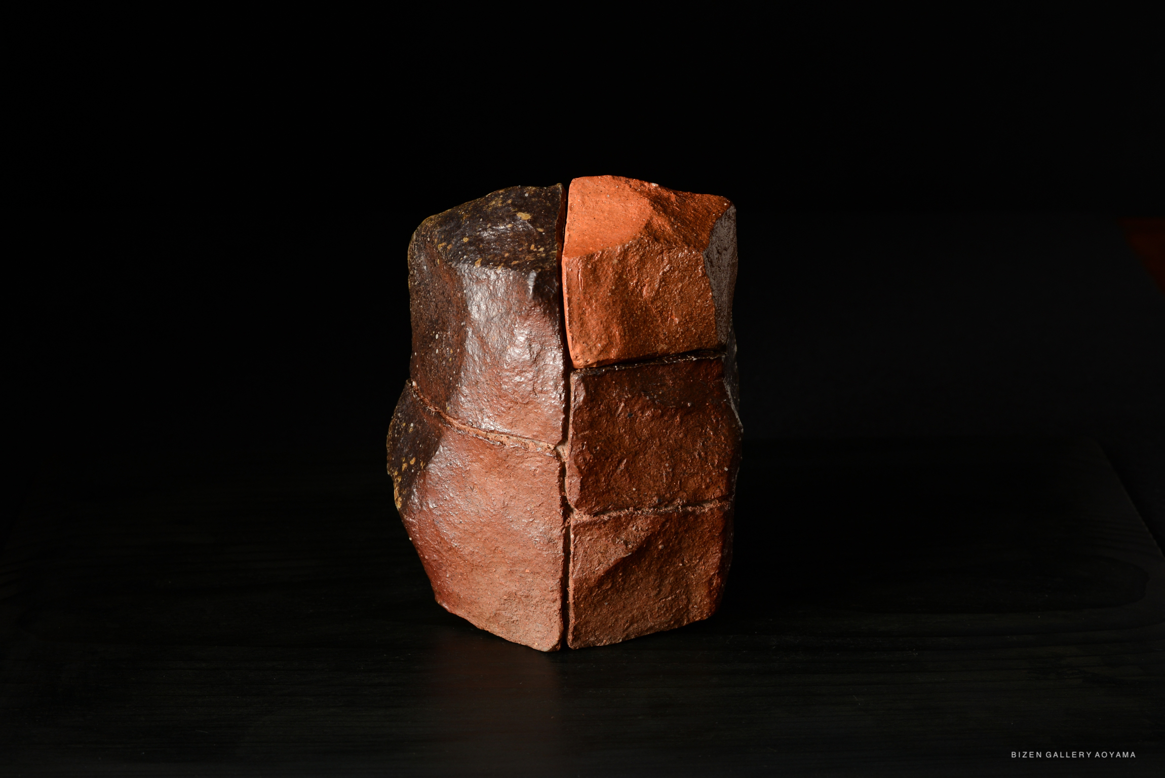 A sculptural piece made of clay, featuring a textured surface with contrasting dark and red sections, set against a black background.