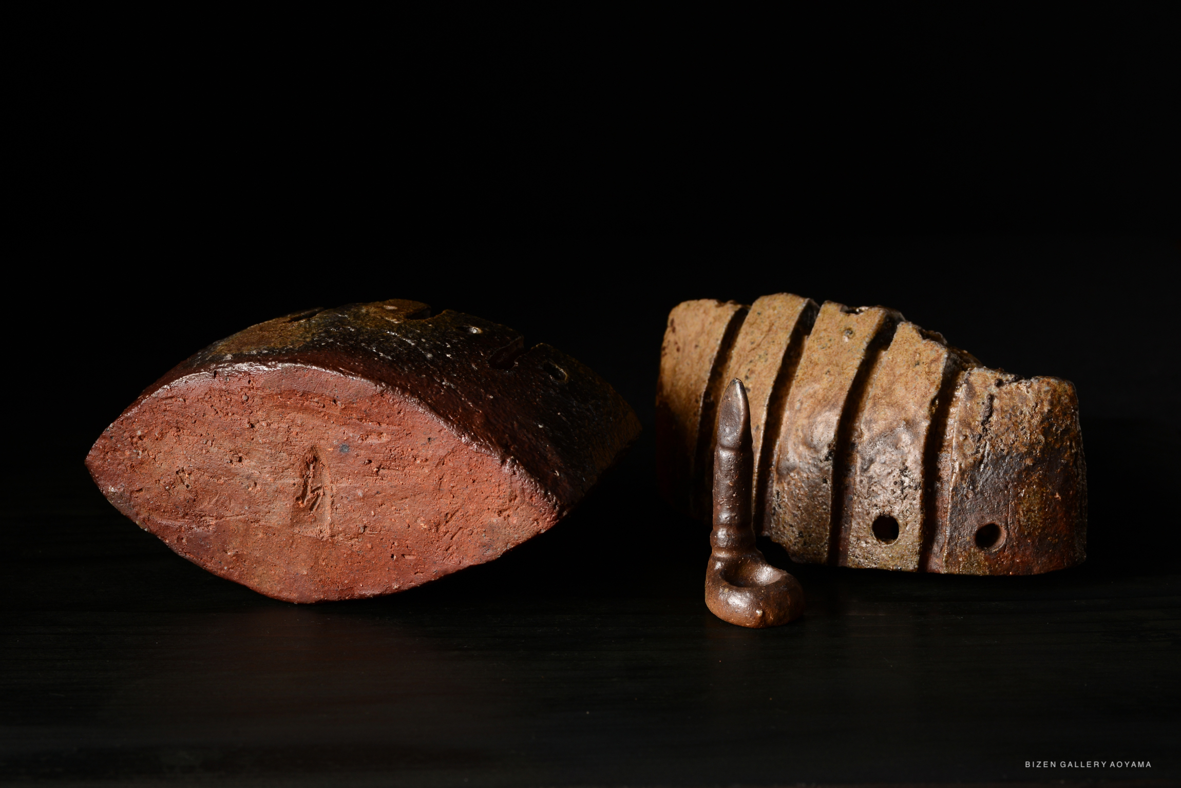 Two pieces of Bizen pottery, featuring a rounded clay form and a flat, textured piece with grooves, along with a small ceramic object resembling a pipe, all set against a dark background.