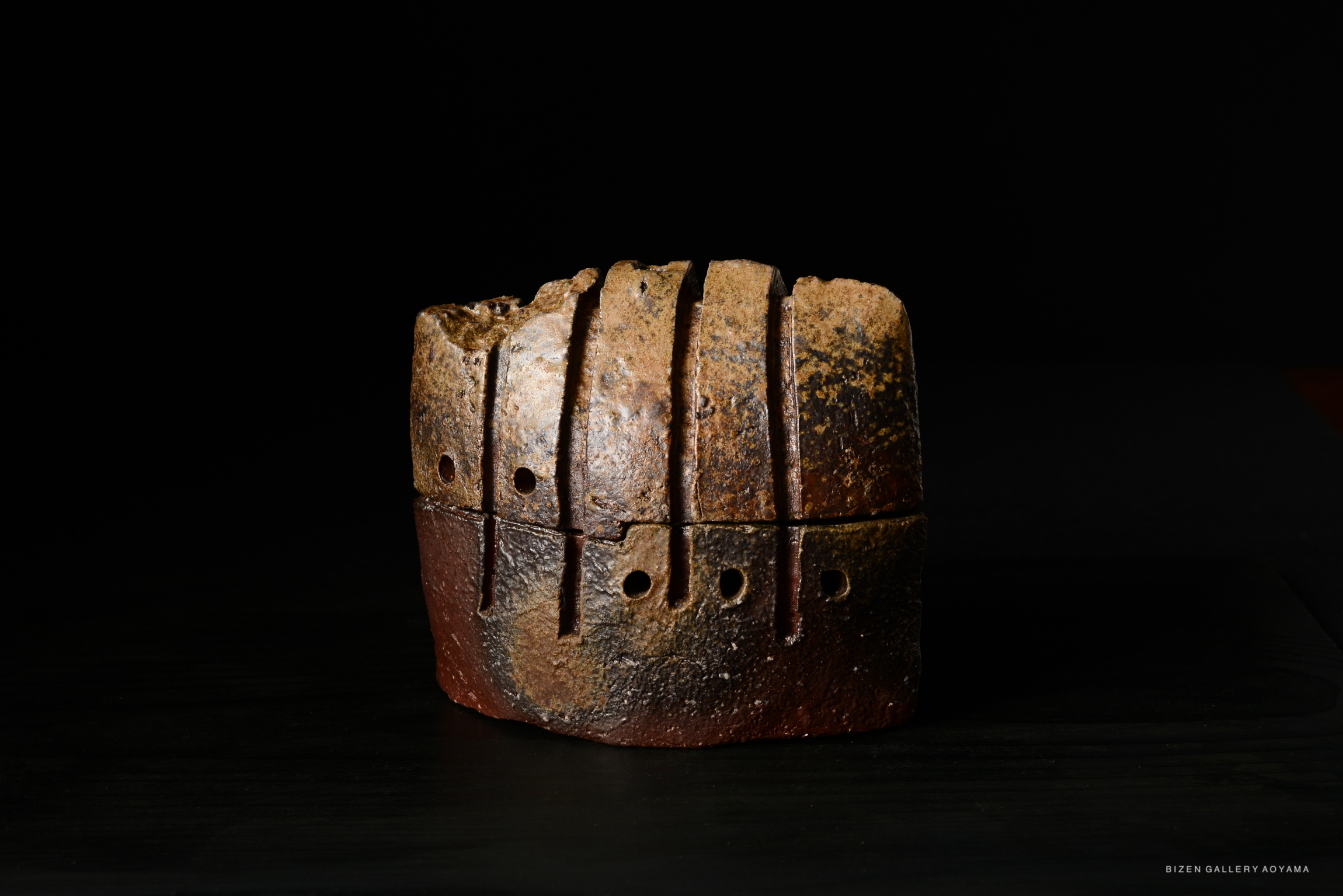A rustic ceramic piece featuring a blocky shape with vertical grooves and small holes, presented against a dark background.
