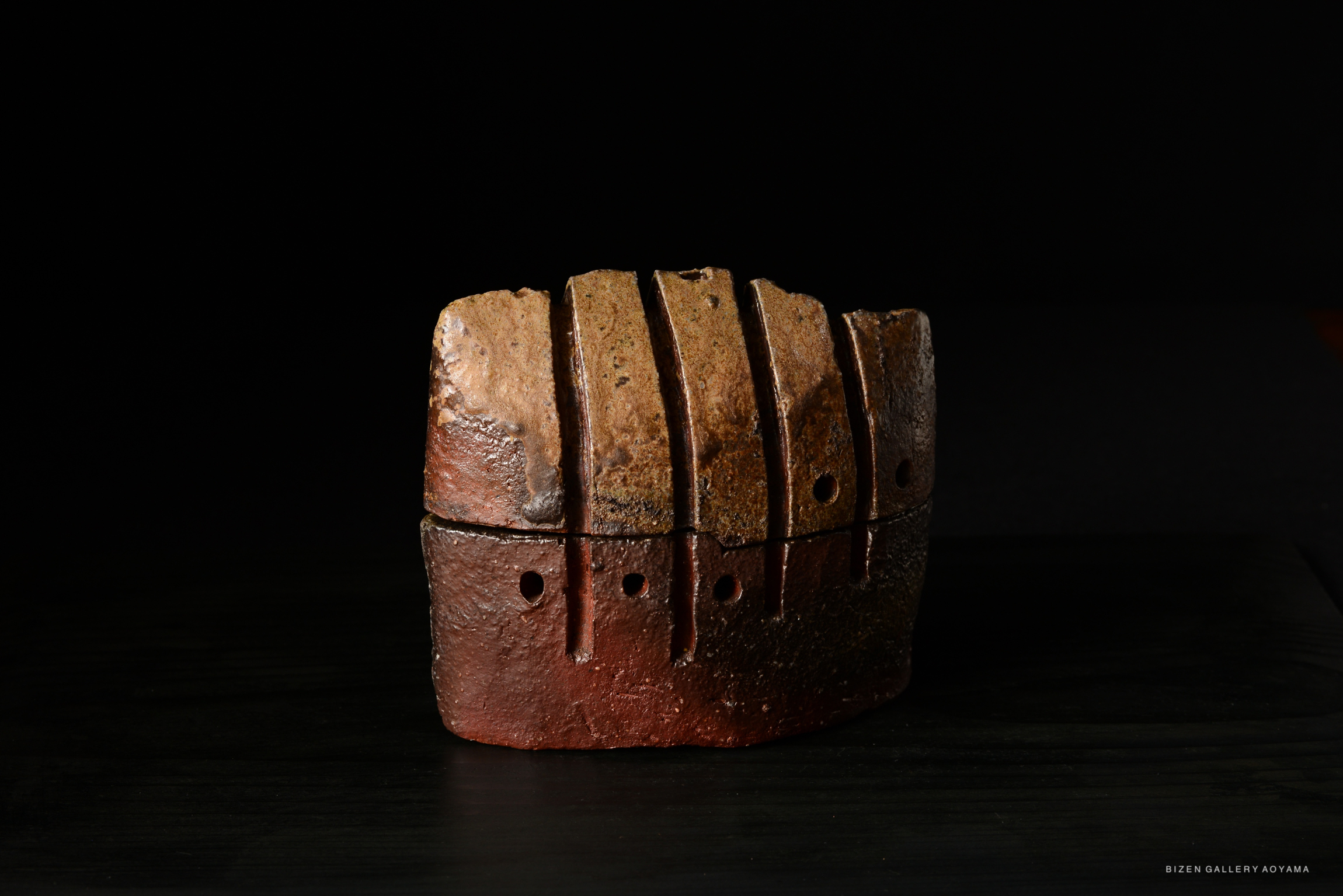 A textured, handcrafted pottery piece with a brown and reddish finish, featuring vertical lines and small holes, set against a dark background.