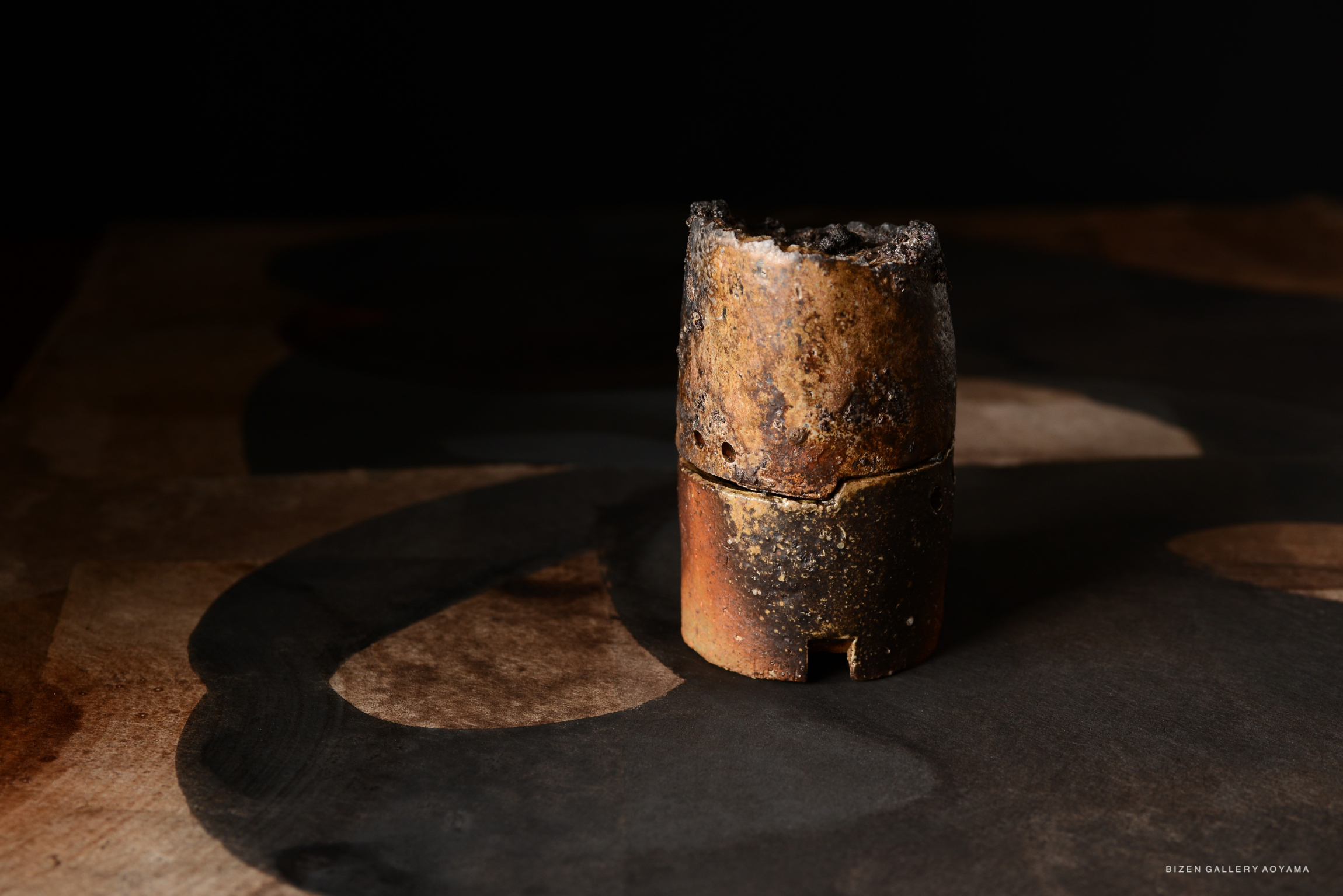 A close-up of a rustic, burnt ceramic piece on a textured surface, featuring dark and earthy tones under soft lighting.