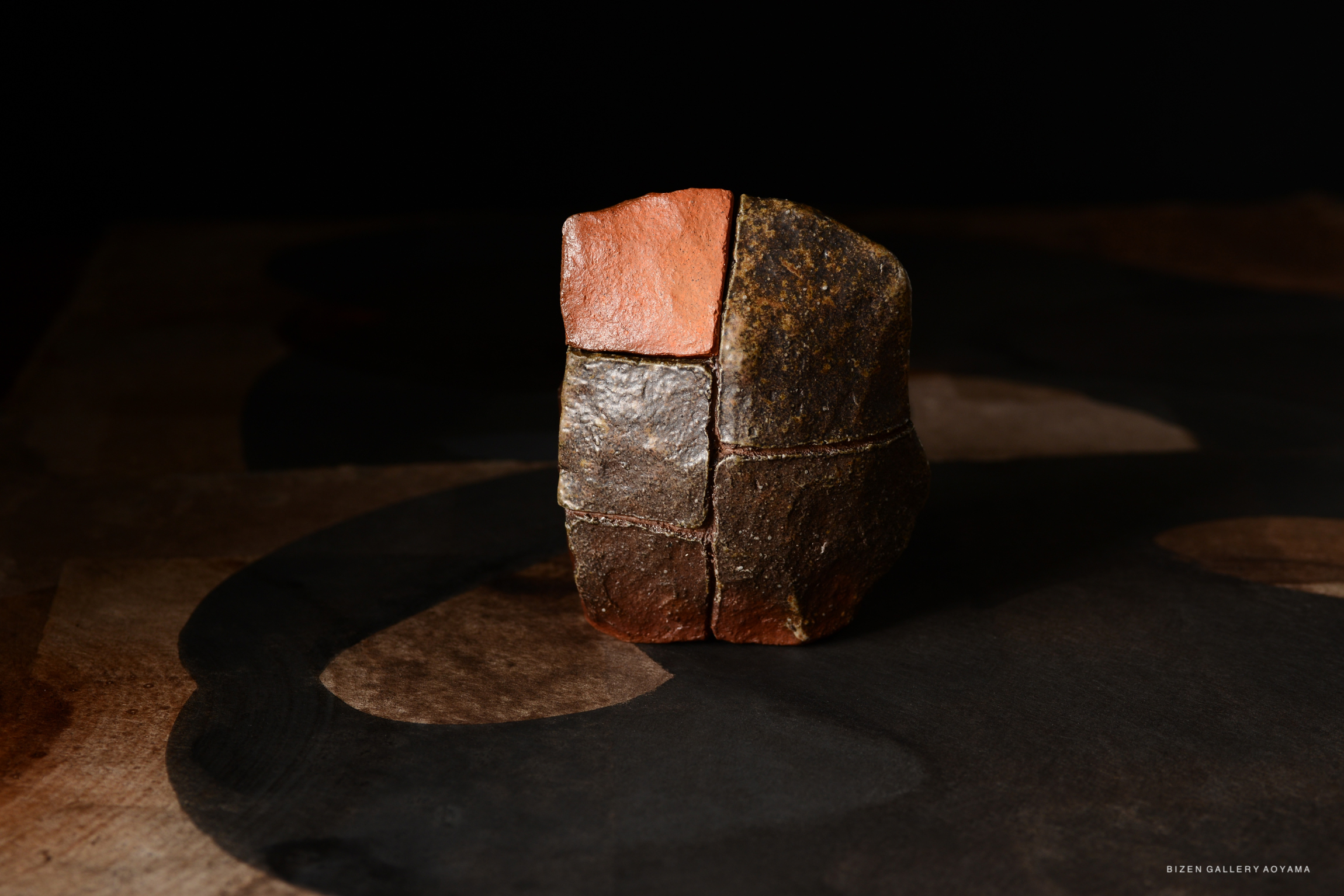 A unique ceramic piece with varied textures and colors, resembling a small sculpture or container, set against a dark background.