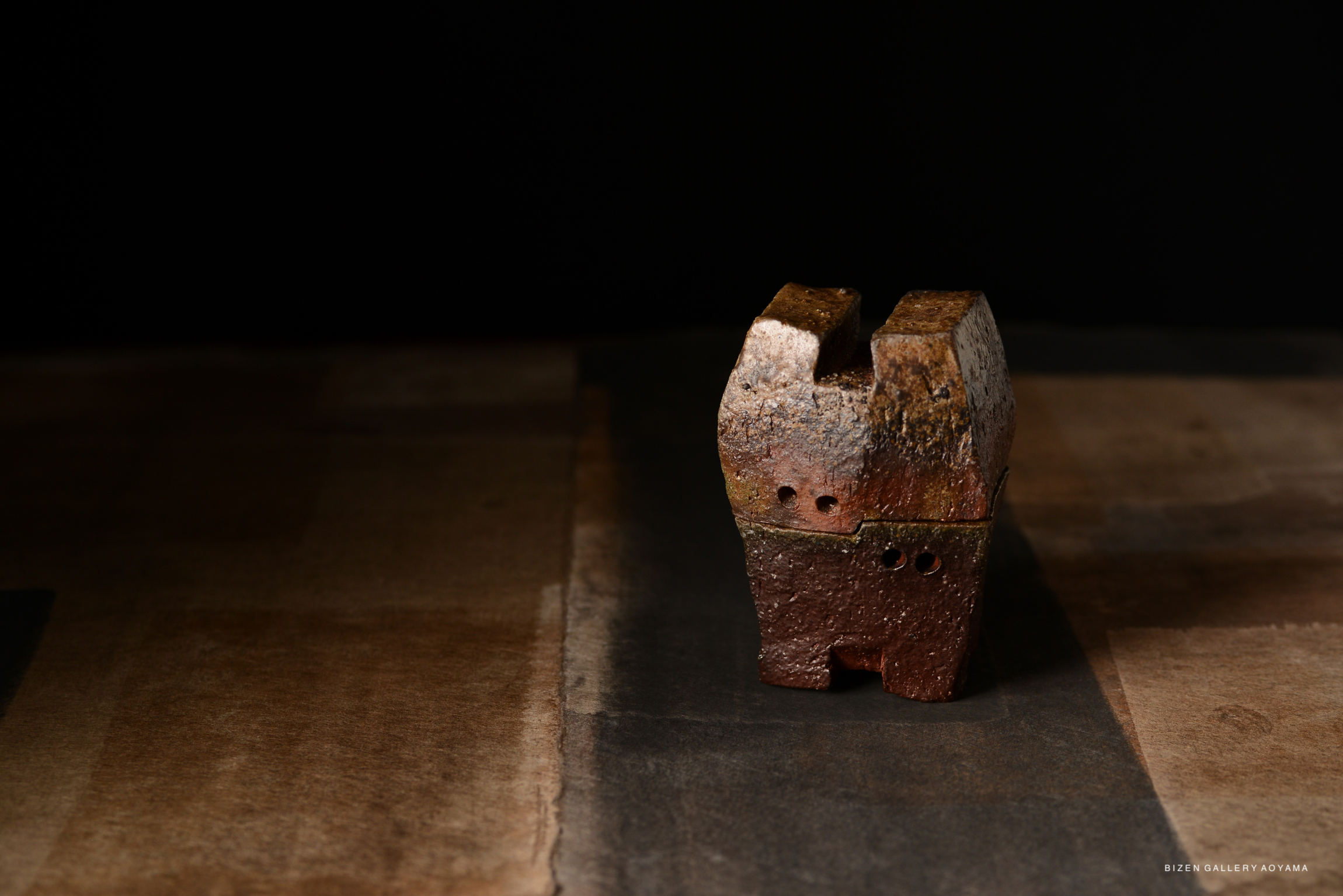 A small, abstract ceramic sculpture with a textured surface, featuring earthy tones of brown and orange, sitting on a wooden surface against a dark background.