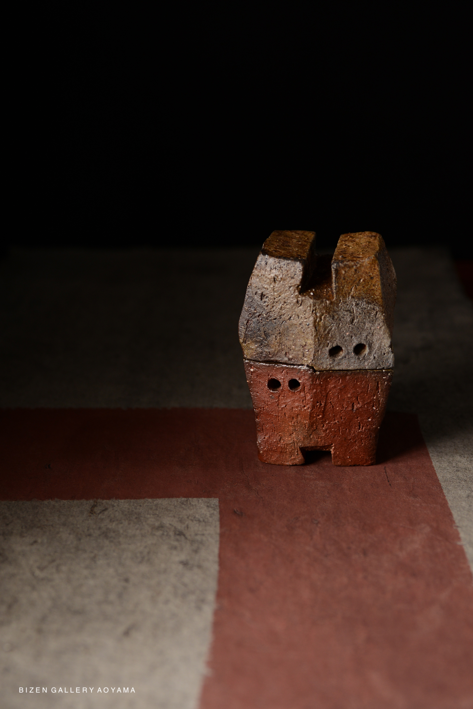 A small, abstract clay sculpture featuring a combination of brown and reddish tones, positioned on a textured surface.