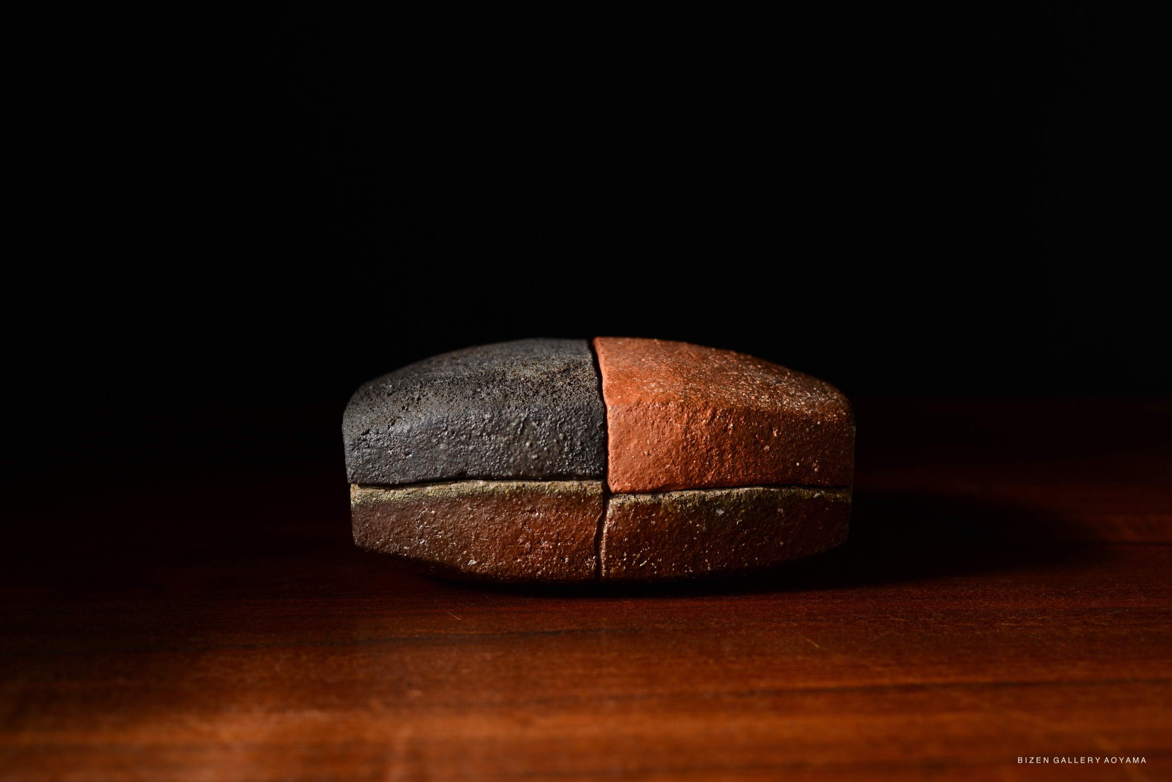 A wooden box containing a ceramic piece with a split design, featuring contrasting earthy tones of dark gray and reddish-brown, placed on a dark surface.
