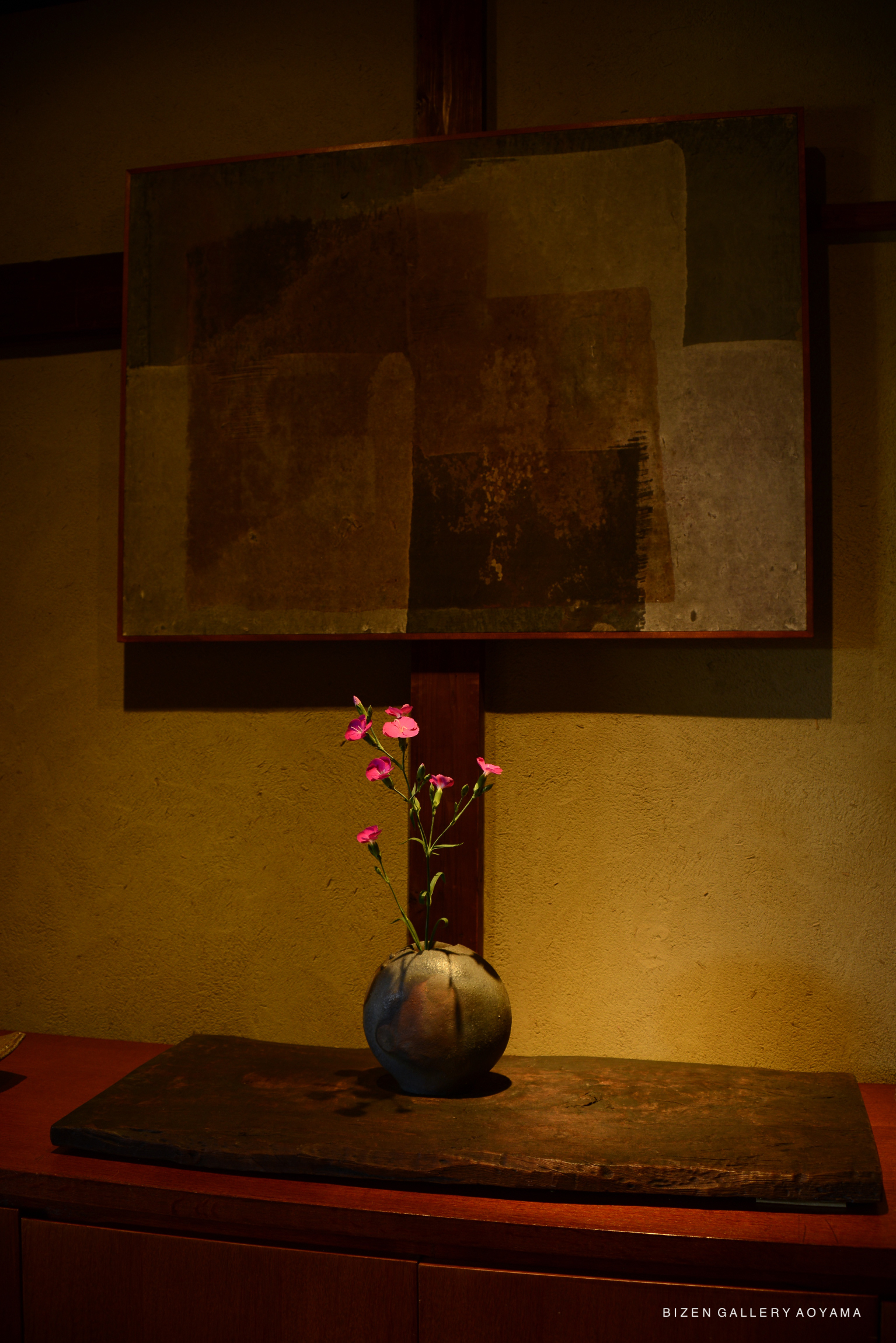 A decorative display featuring a round Bizen pottery vase holding pink flowers on a wooden table, with an abstract painting in the background.