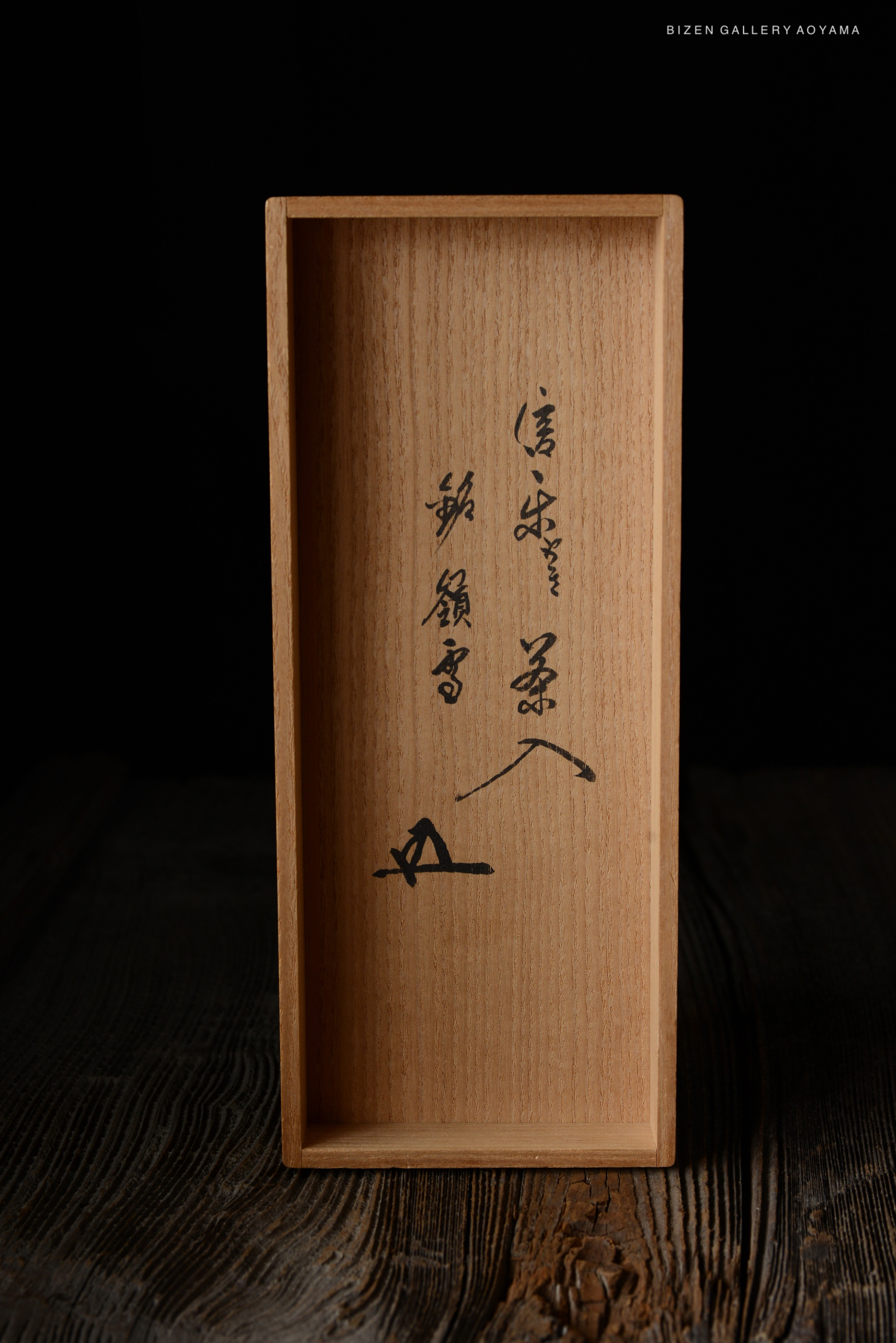 A wooden box with engraved calligraphy, likely used for a Shigaraki Chaire, resting on a rustic wooden surface.