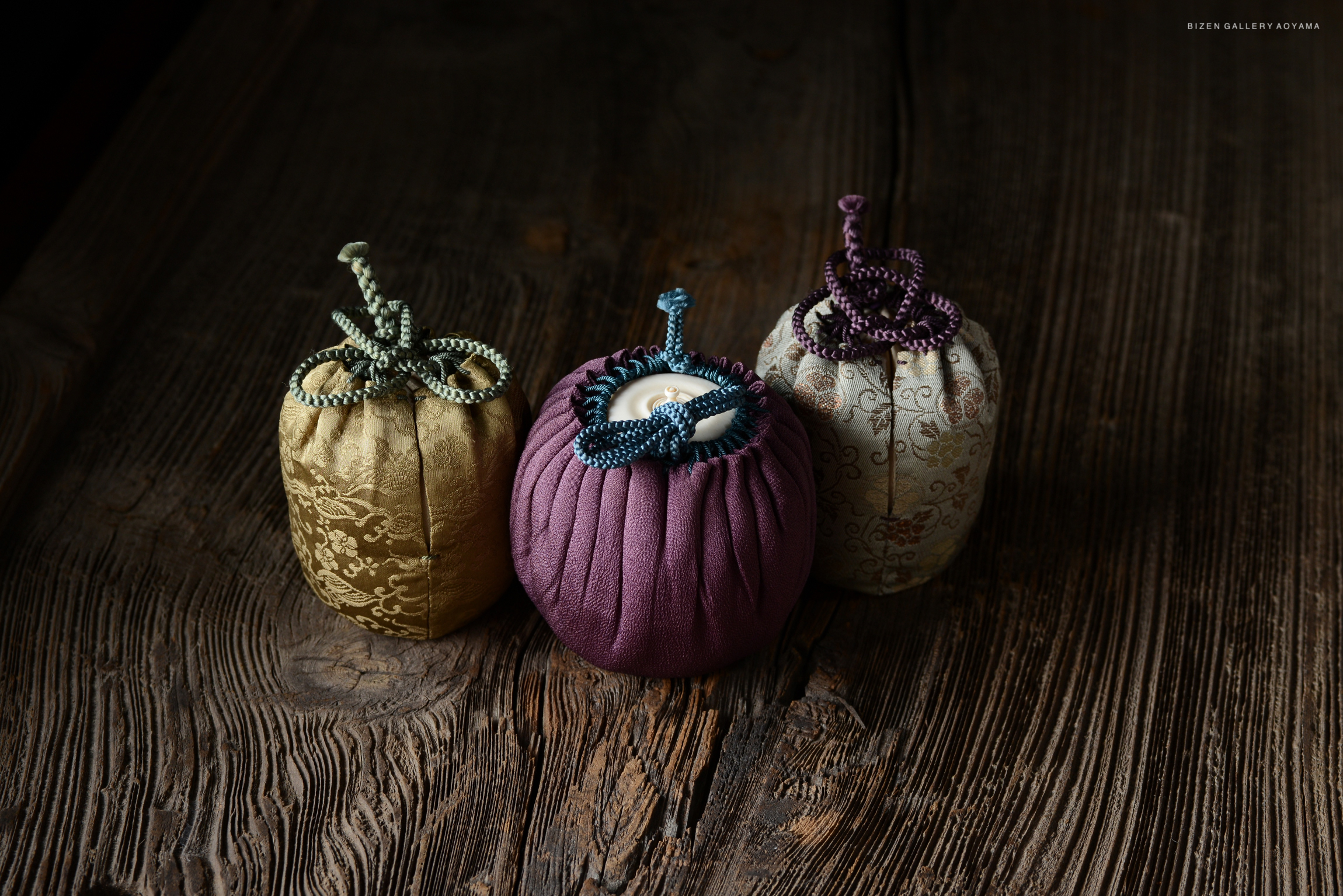 Three traditional Japanese tea caddies in decorative fabrics, featuring knots and intricate designs, displayed on a rustic wooden surface.