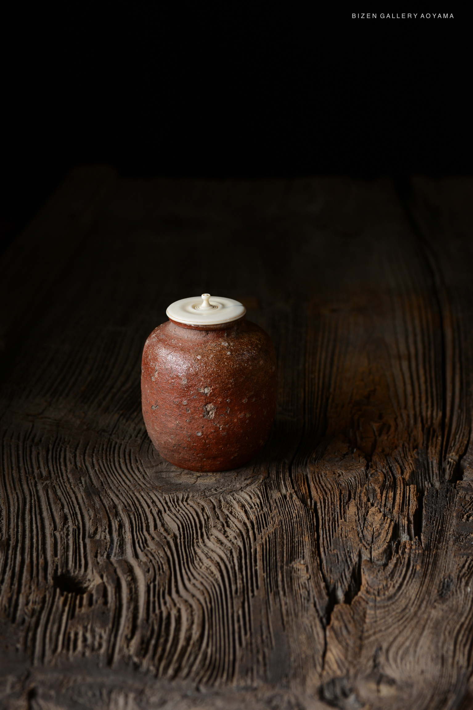 A Shigaraki Chaire tea storage container, featuring a rustic glazed finish and a light-colored lid, displayed on a textured wooden surface.